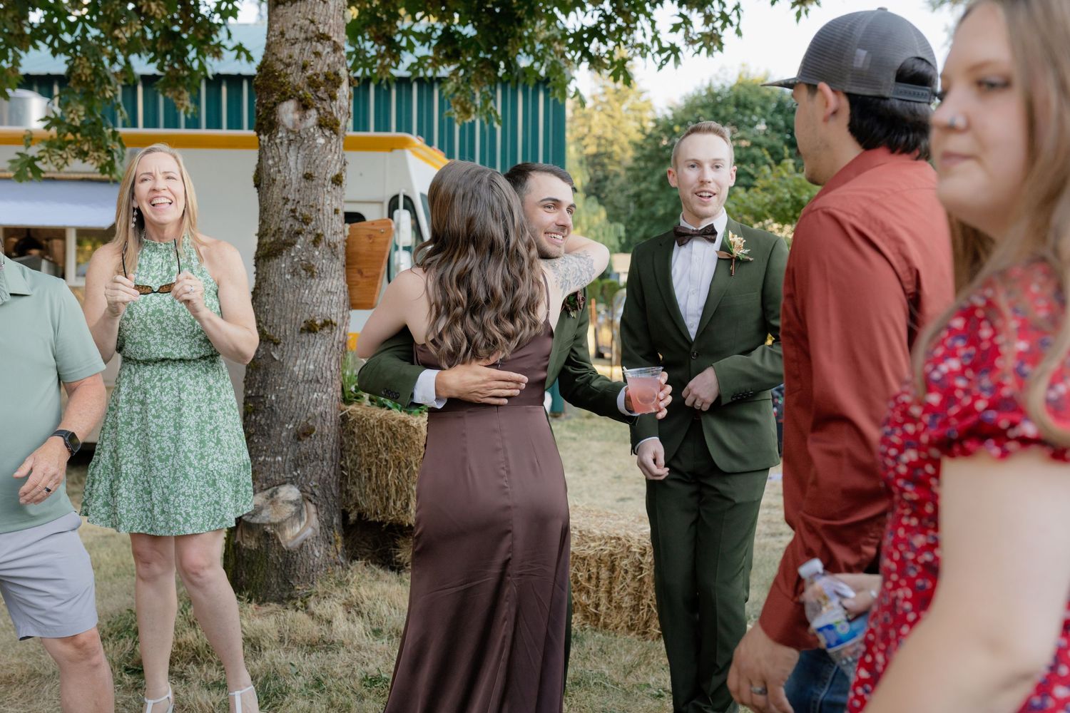 Wedding guests mingling outdoors near hay bales, groom in green suit holding champagne with bride.