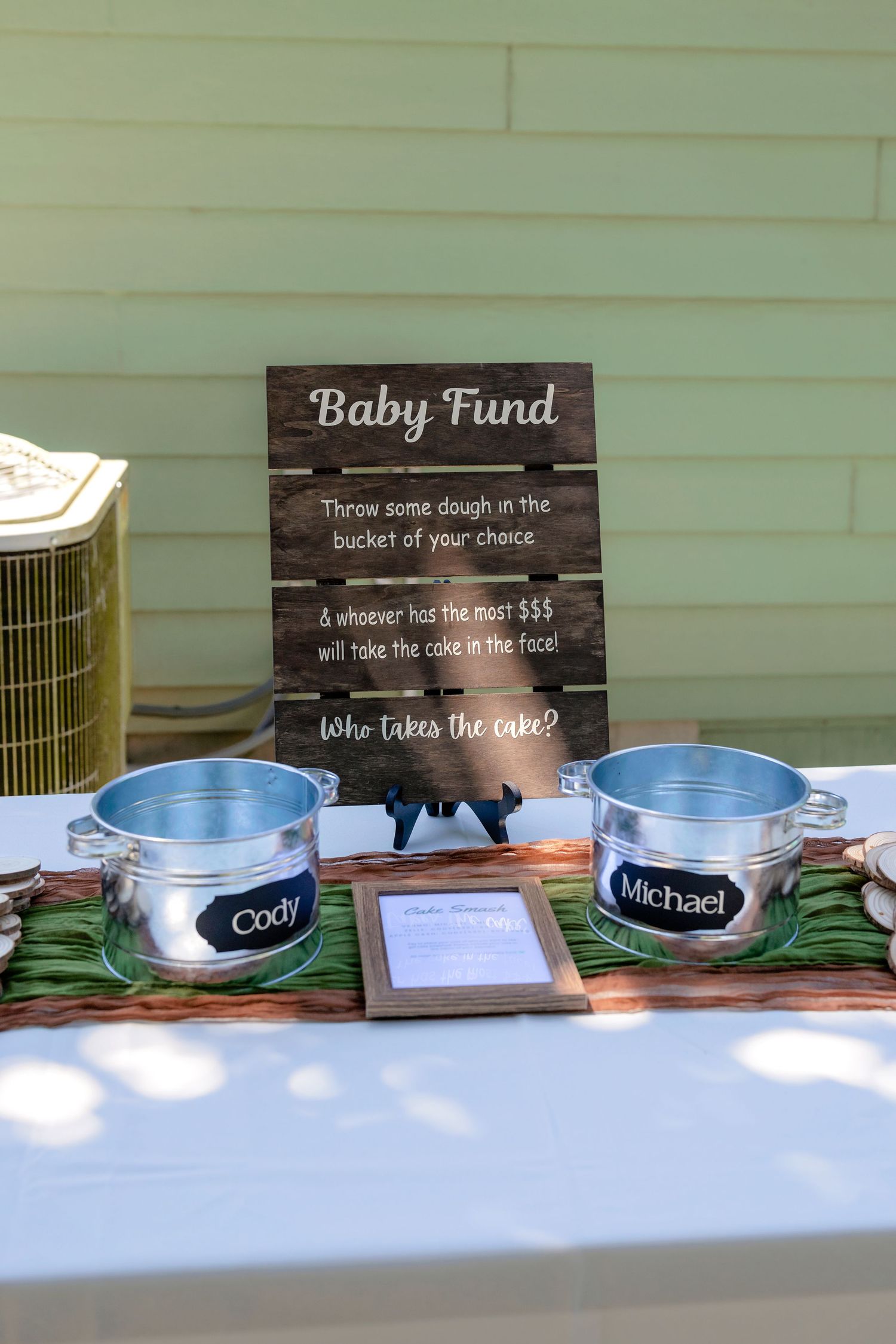 Baby fund donation buckets labeled Cody and Michael at a rustic outdoor baby shower with a wooden sign.
