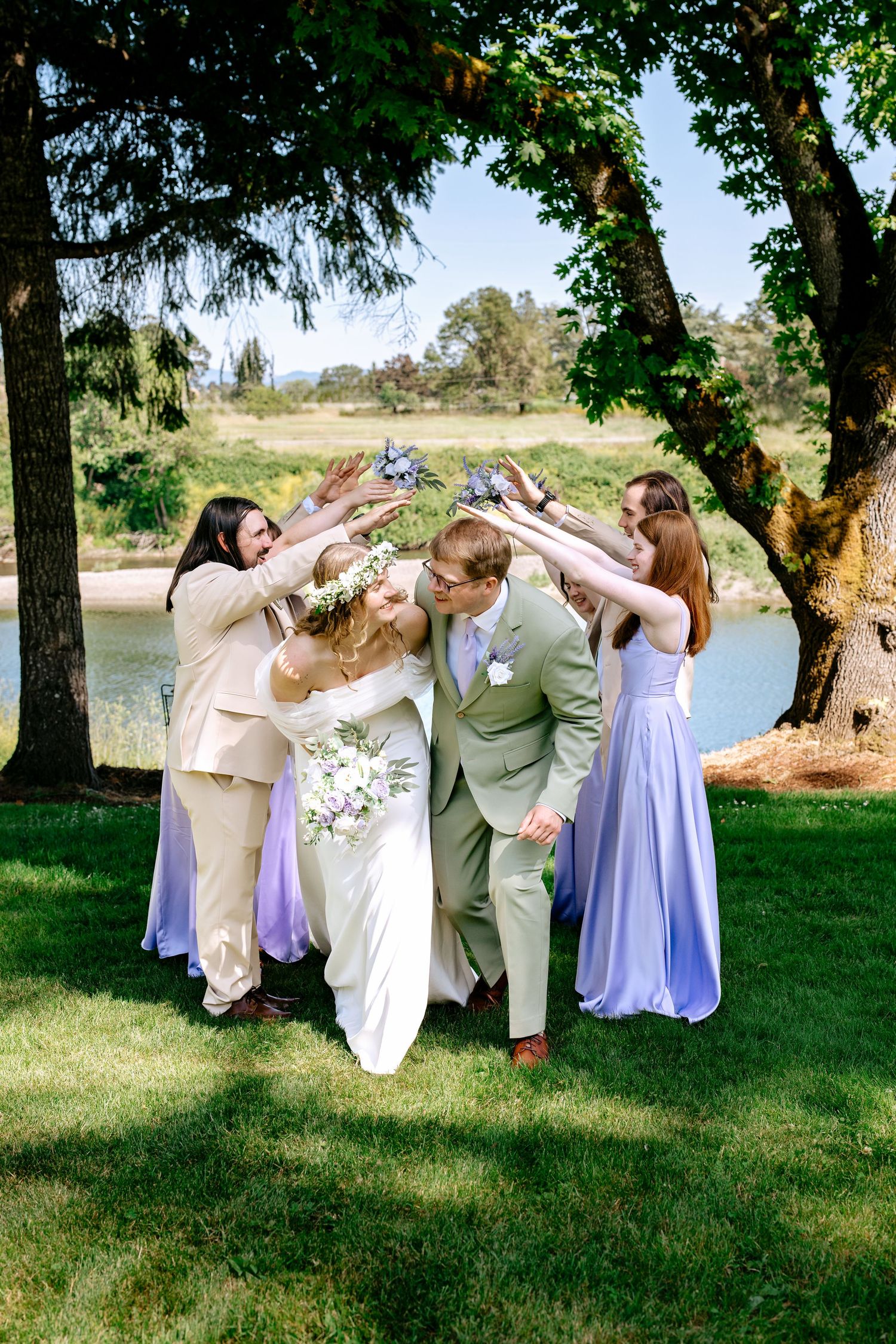 Wedding party celebrating newlyweds with raised bouquets arch while couple kisses under trees near a lake.