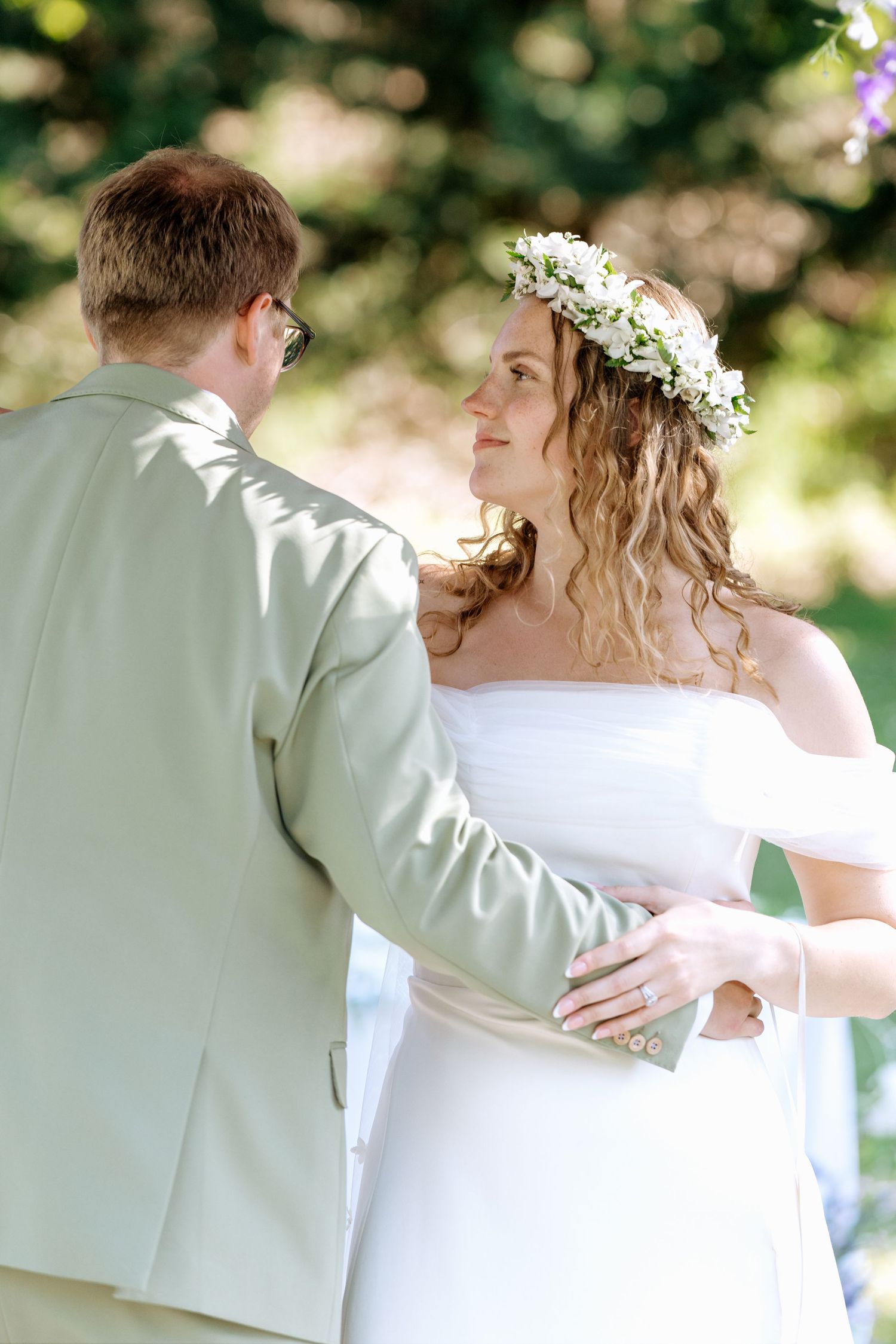 Bride wearing floral crown gazes lovingly at groom during outdoor wedding ceremony in sunlit garden setting.