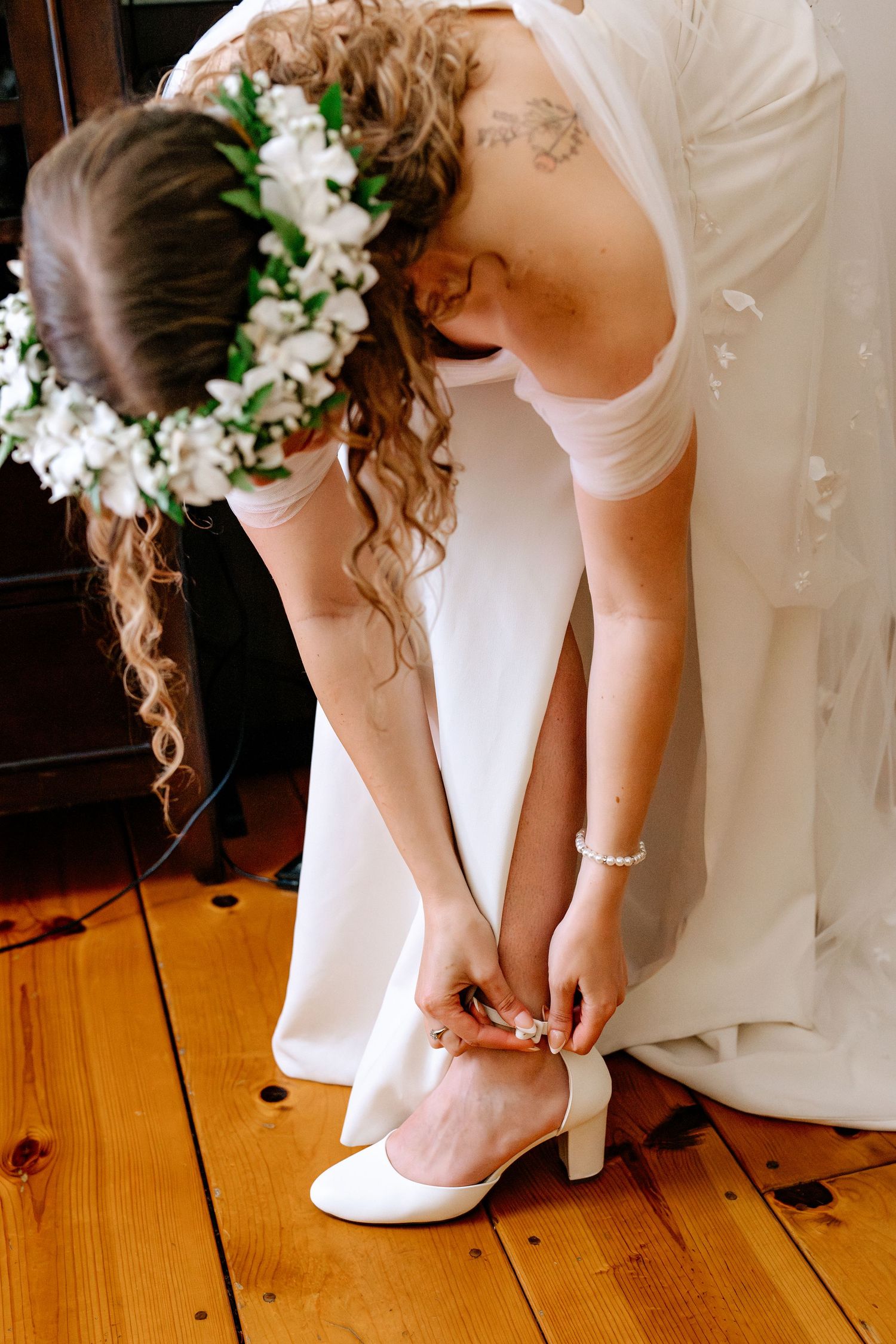 Bride in white gown buckles her white heeled shoe on a wooden floor, floral crown in her curly hair.