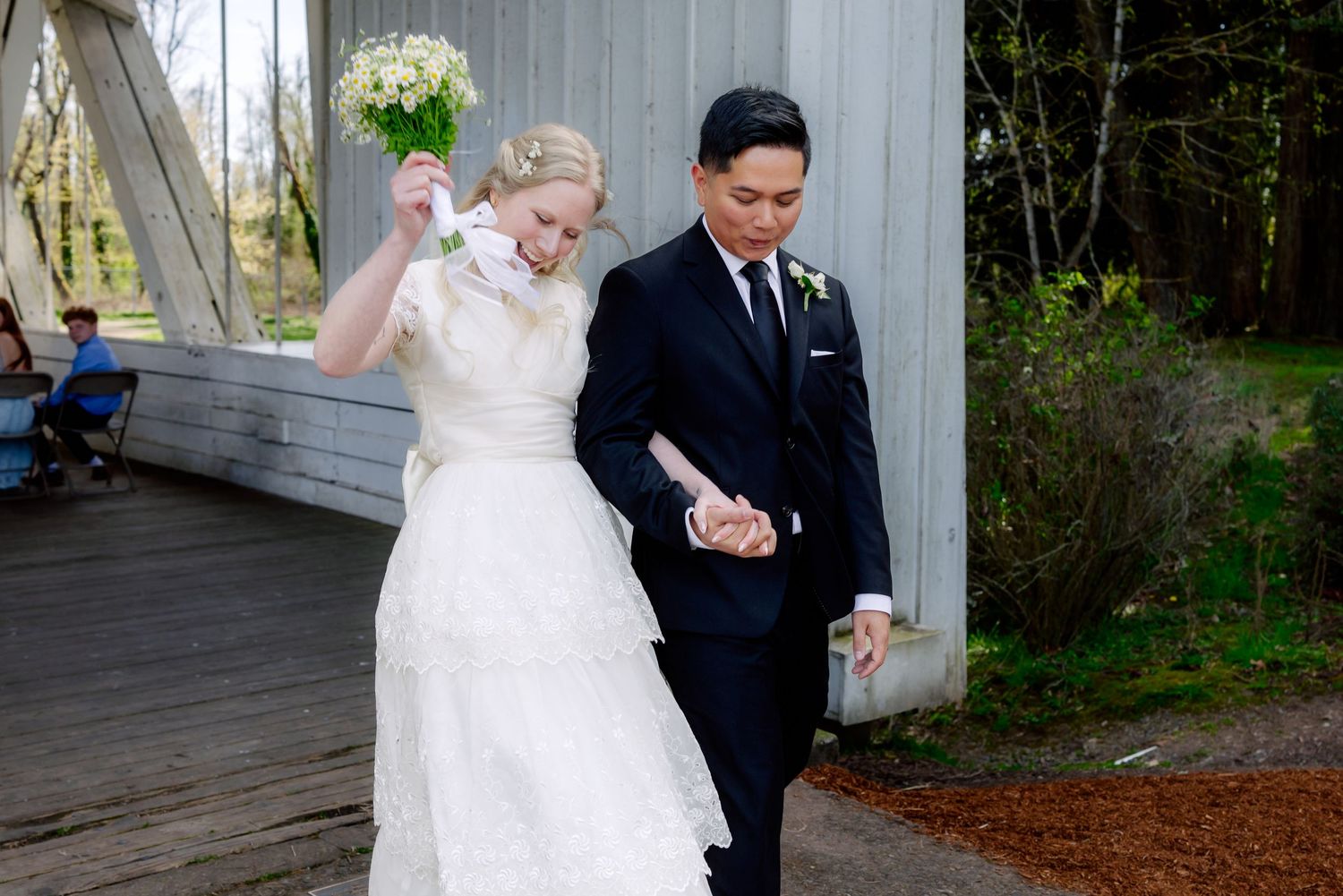 A bride in white gown holding bouquet walks with groom in black suit at outdoor wedding venue.
