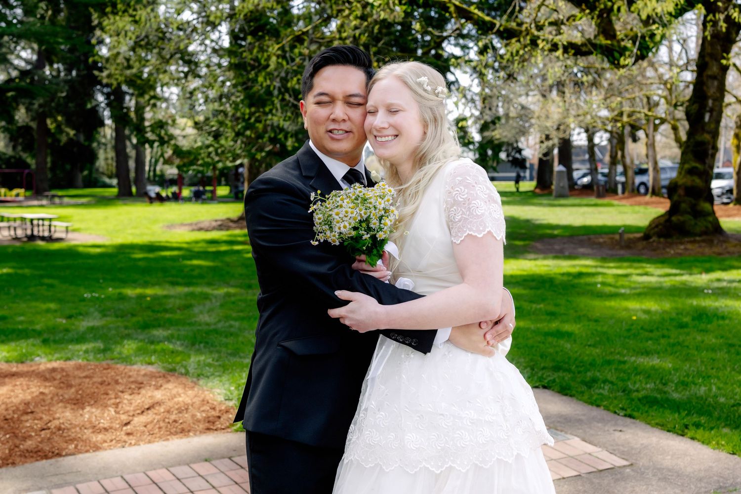A groom in a black suit embraces his bride in a white lace gown holding a bouquet in a lush green park.