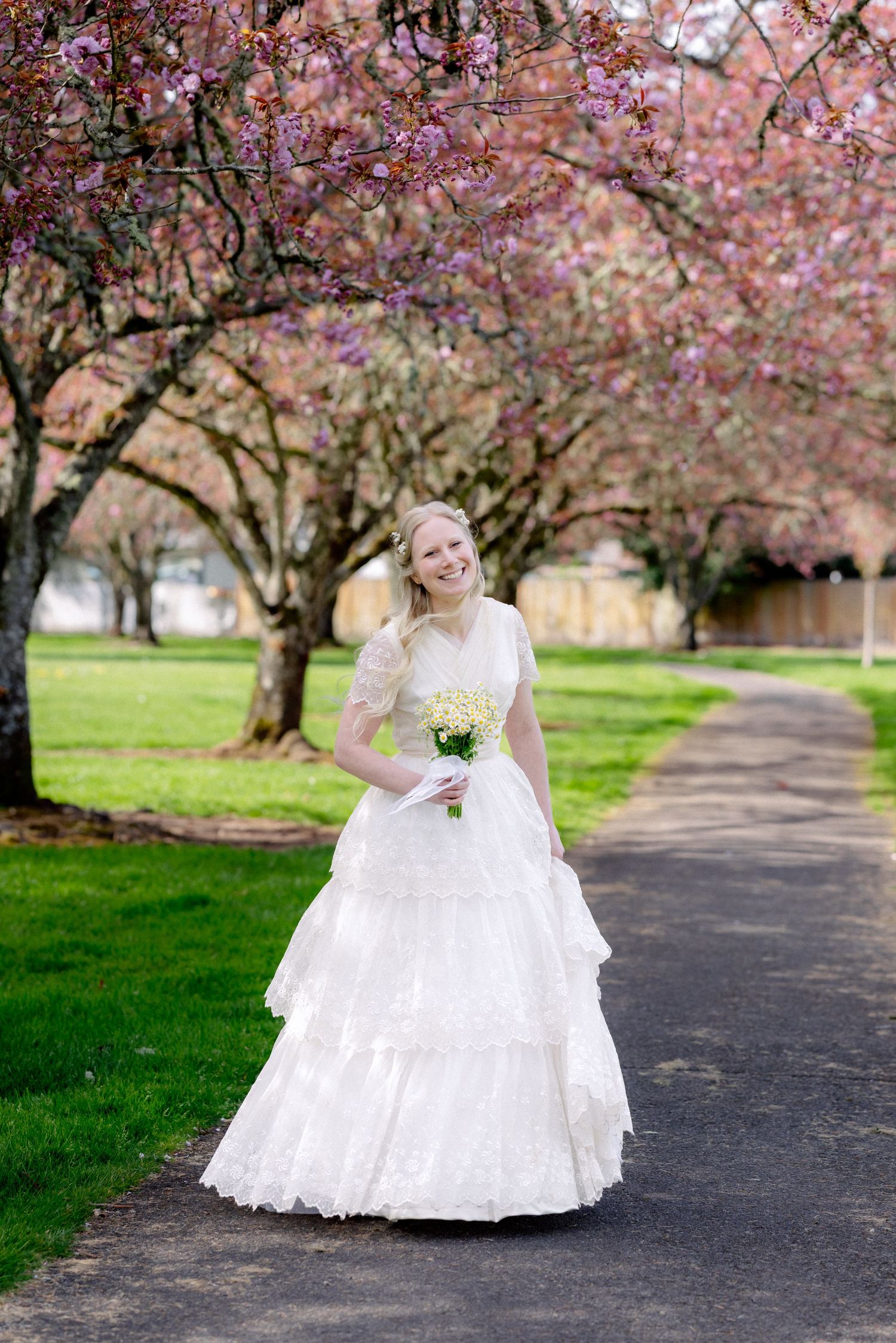 Smiling bride in white lace gown holding bouquet under blooming pink cherry blossom trees.