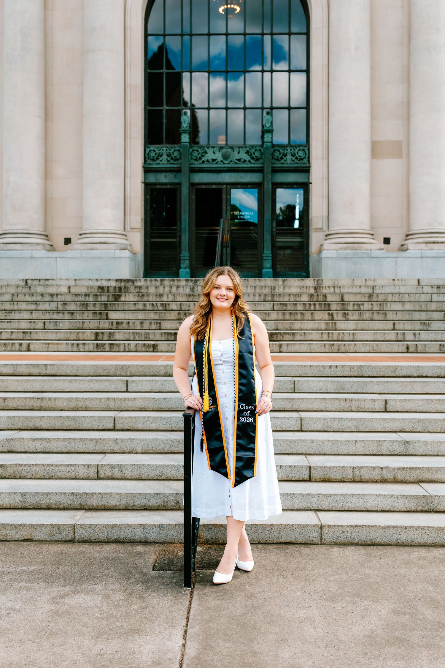 A smiling graduate poses in a white dress with a black and gold stole on stone steps outside a classical building.