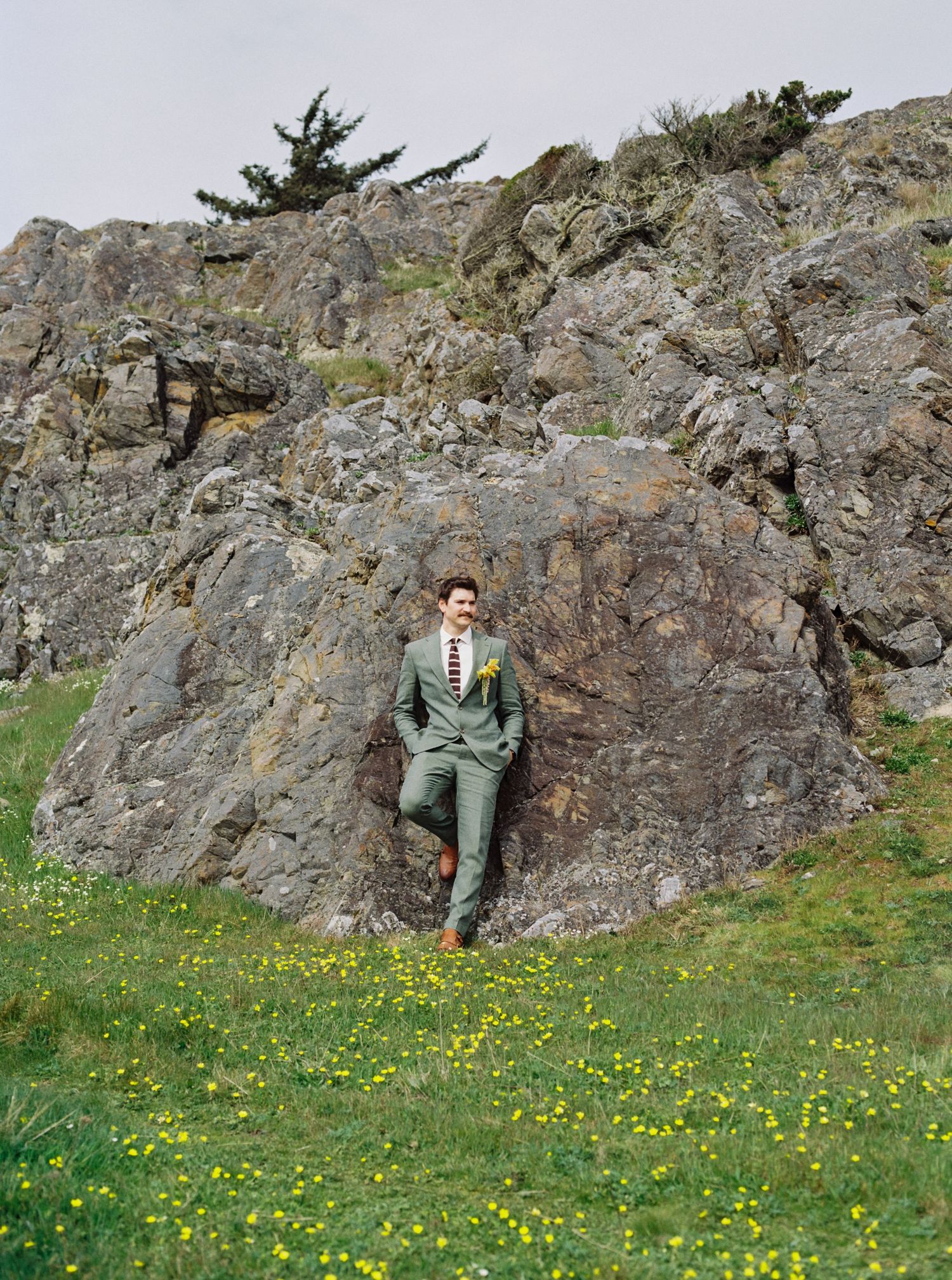 Groom in gray suit leans against rugged rock face surrounded by yellow wildflowers in green meadow.
