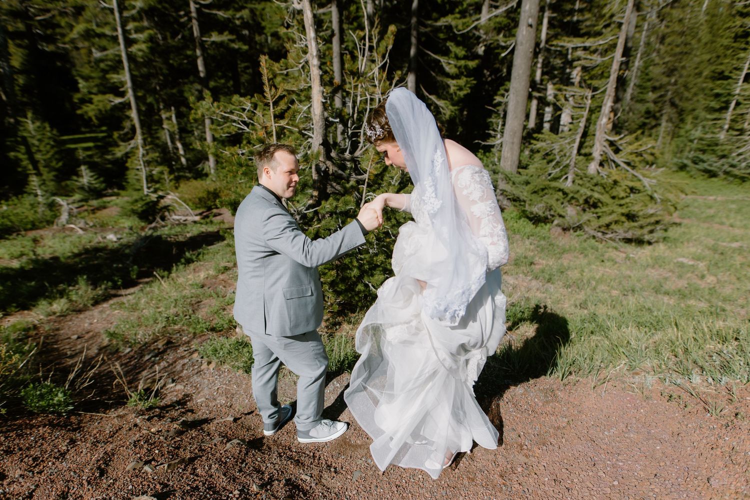 Groom in gray suit helps bride in lace gown navigate forest trail on wedding day.