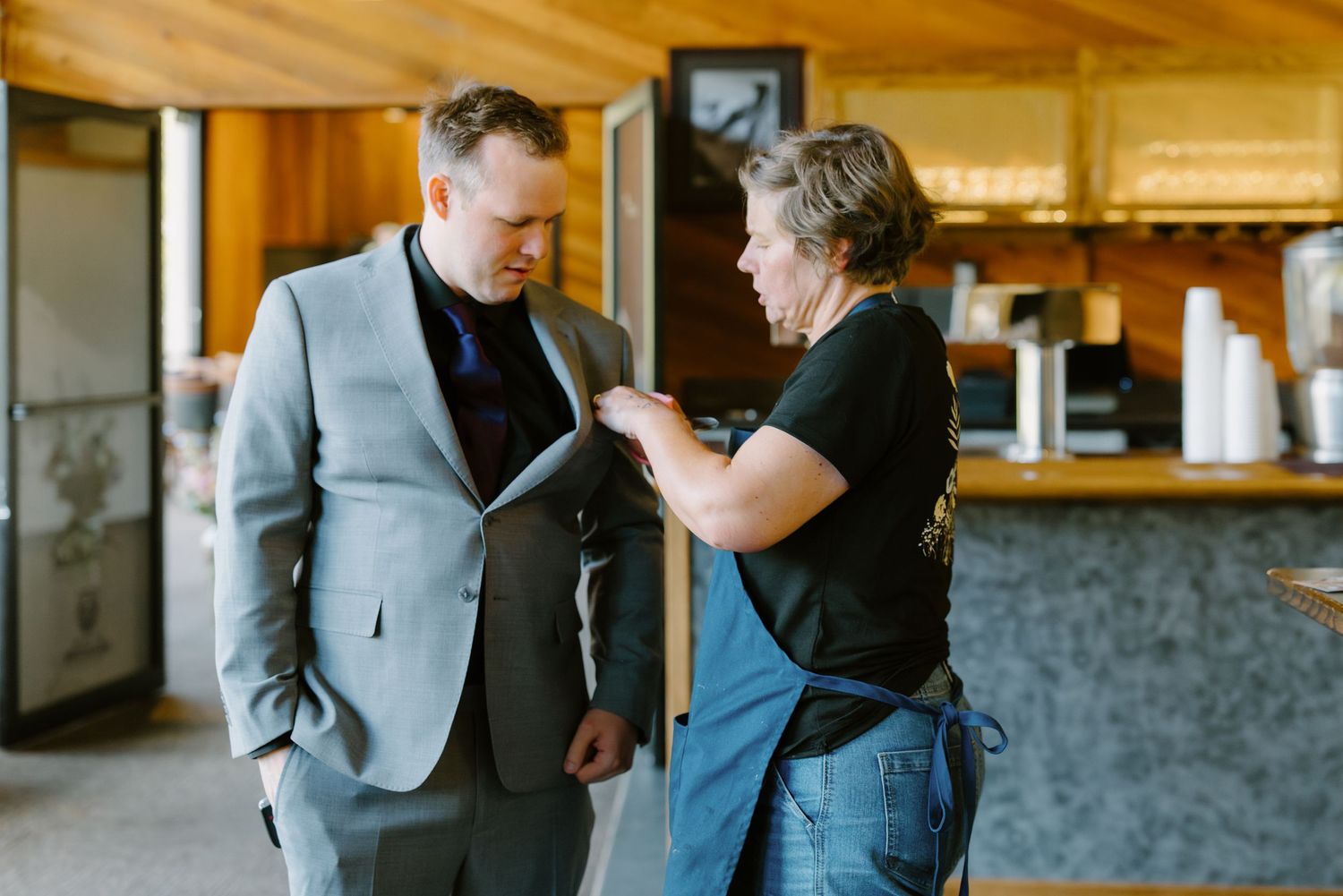 Man in gray suit reviews document with woman in apron at rustic wooden venue counter.