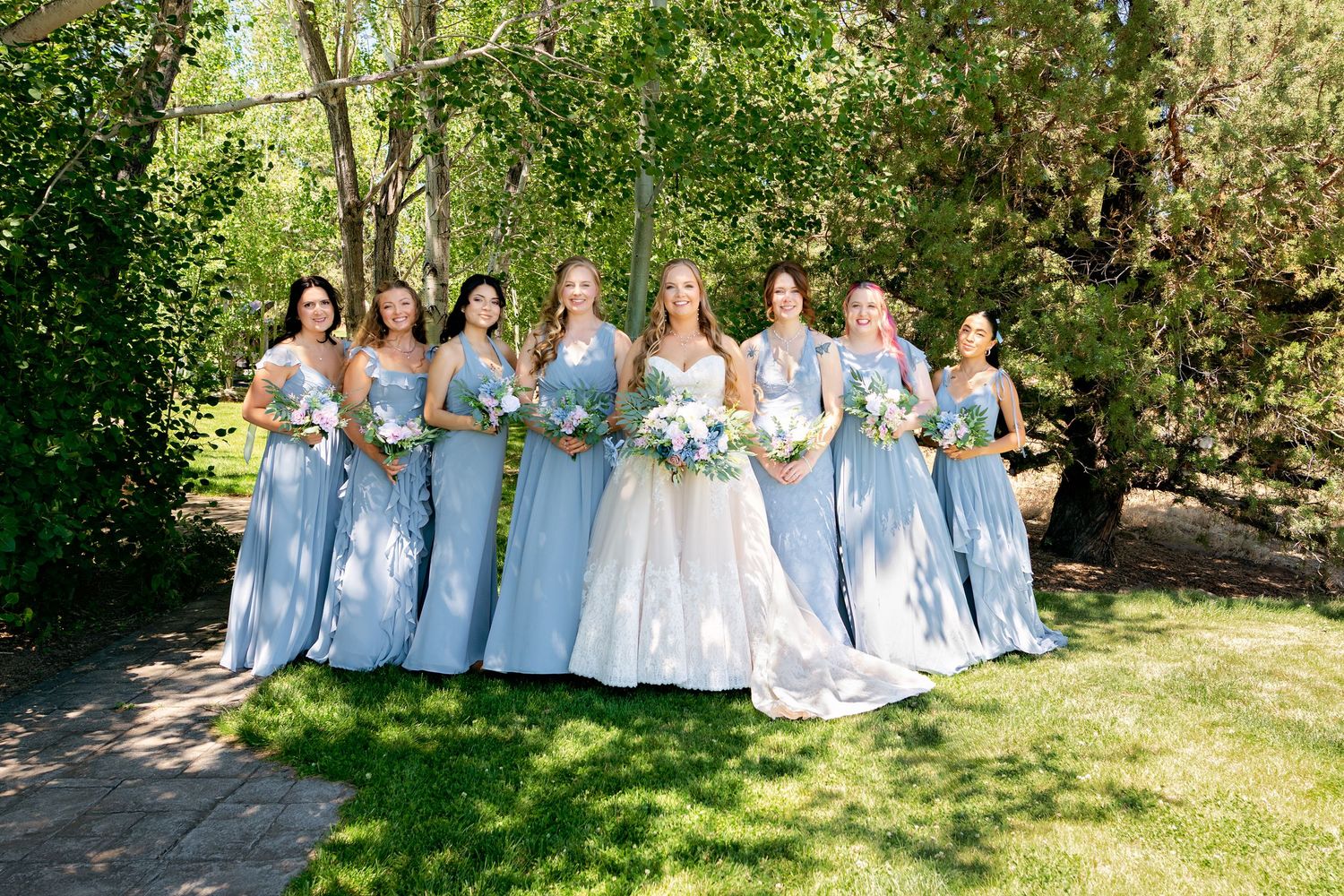 Bride poses with seven bridesmaids in matching light blue gowns holding white bouquets outdoors.