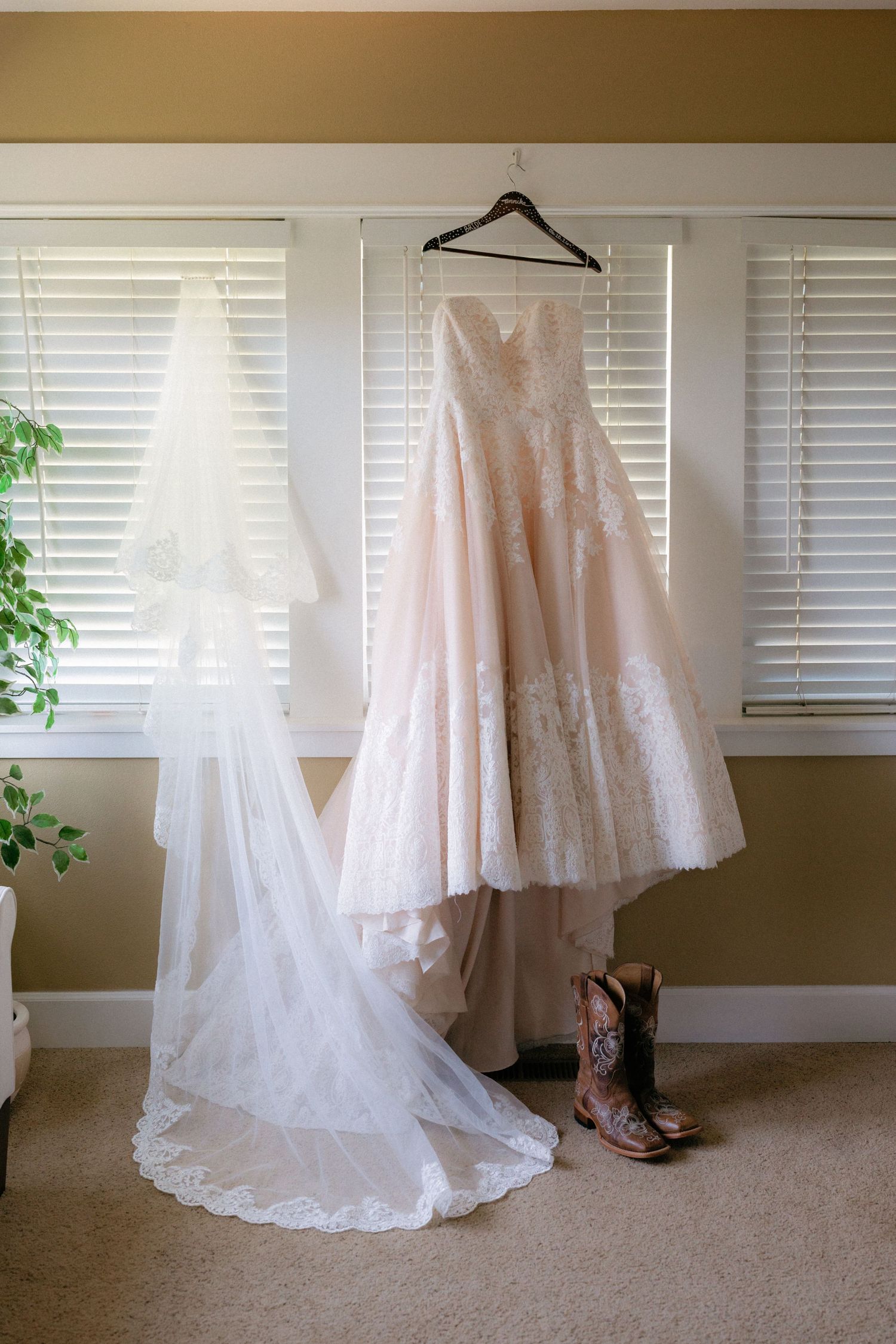 Elegant lace wedding dress hanging with a veil and cowboy boots below, displayed against window blinds.