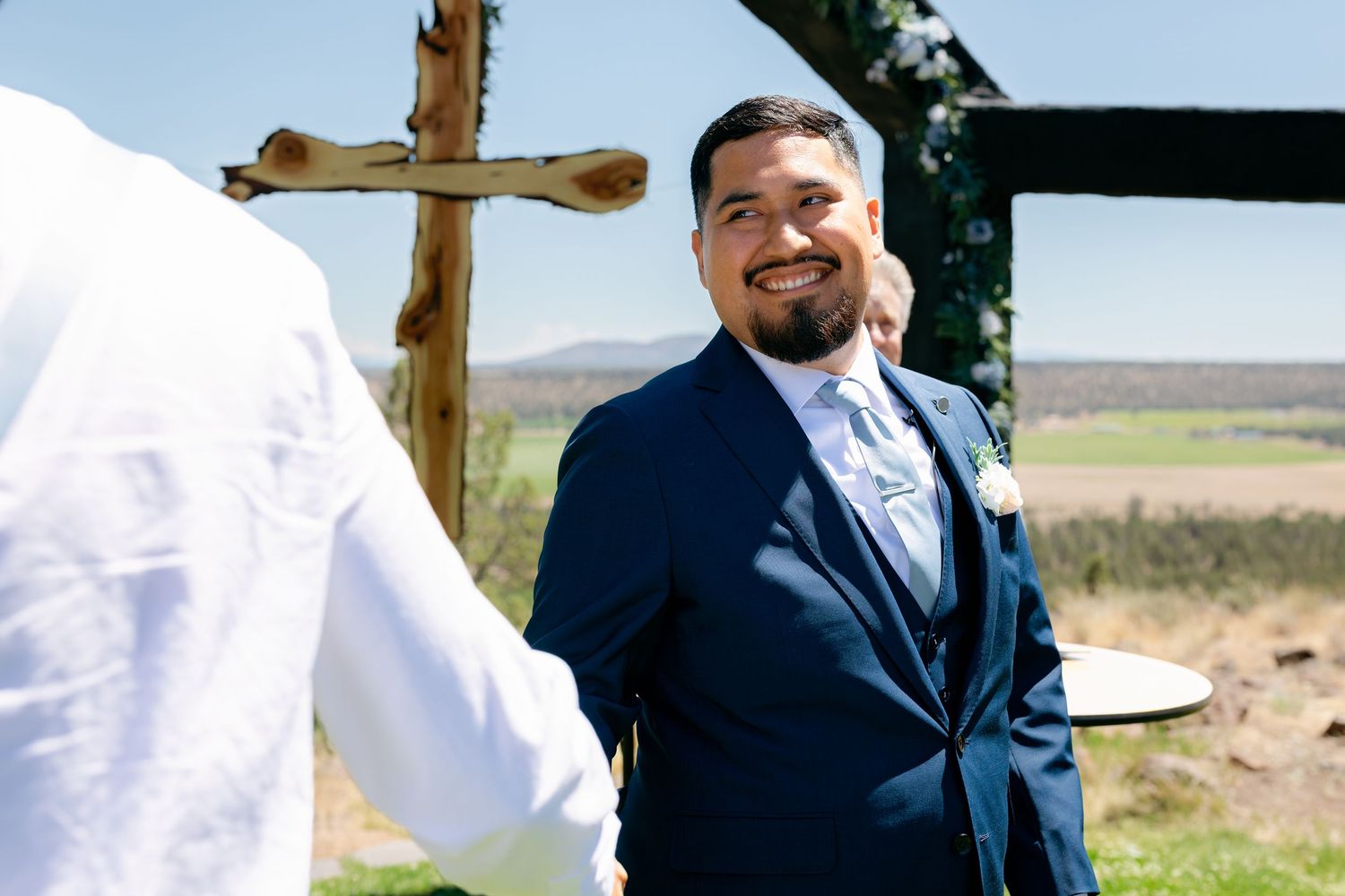 Smiling groom in navy suit shaking hands at outdoor wedding ceremony with wooden cross in background.