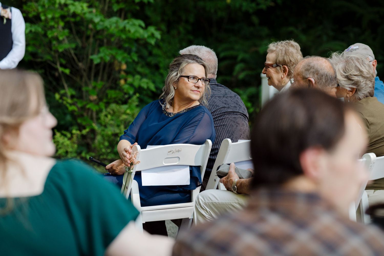 Outdoor gathering with guests seated in white chairs surrounded by lush green trees at a daytime event.