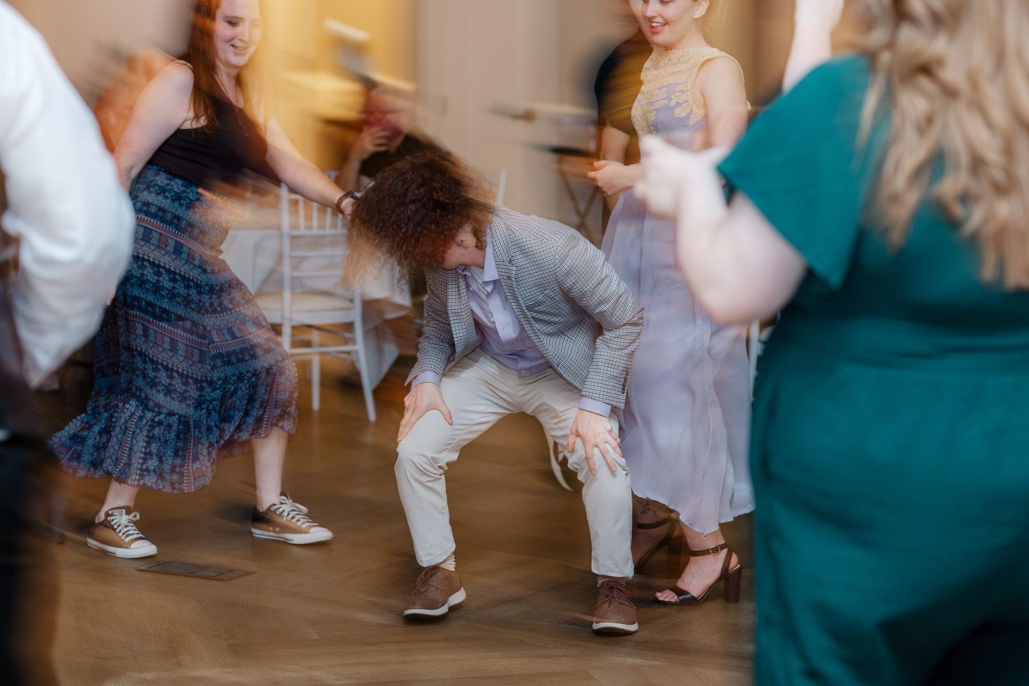 Guests dancing energetically at a wedding reception in a bright venue, man in gray suit doing low dance move.