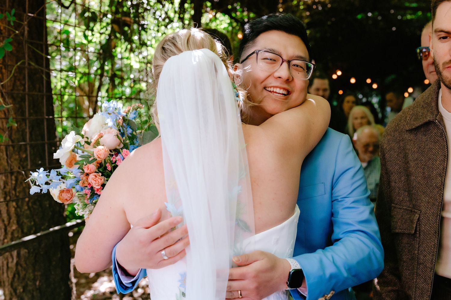 Smiling groom in blue suit hugging a guest while bride holds colorful bouquet at wedding celebration.