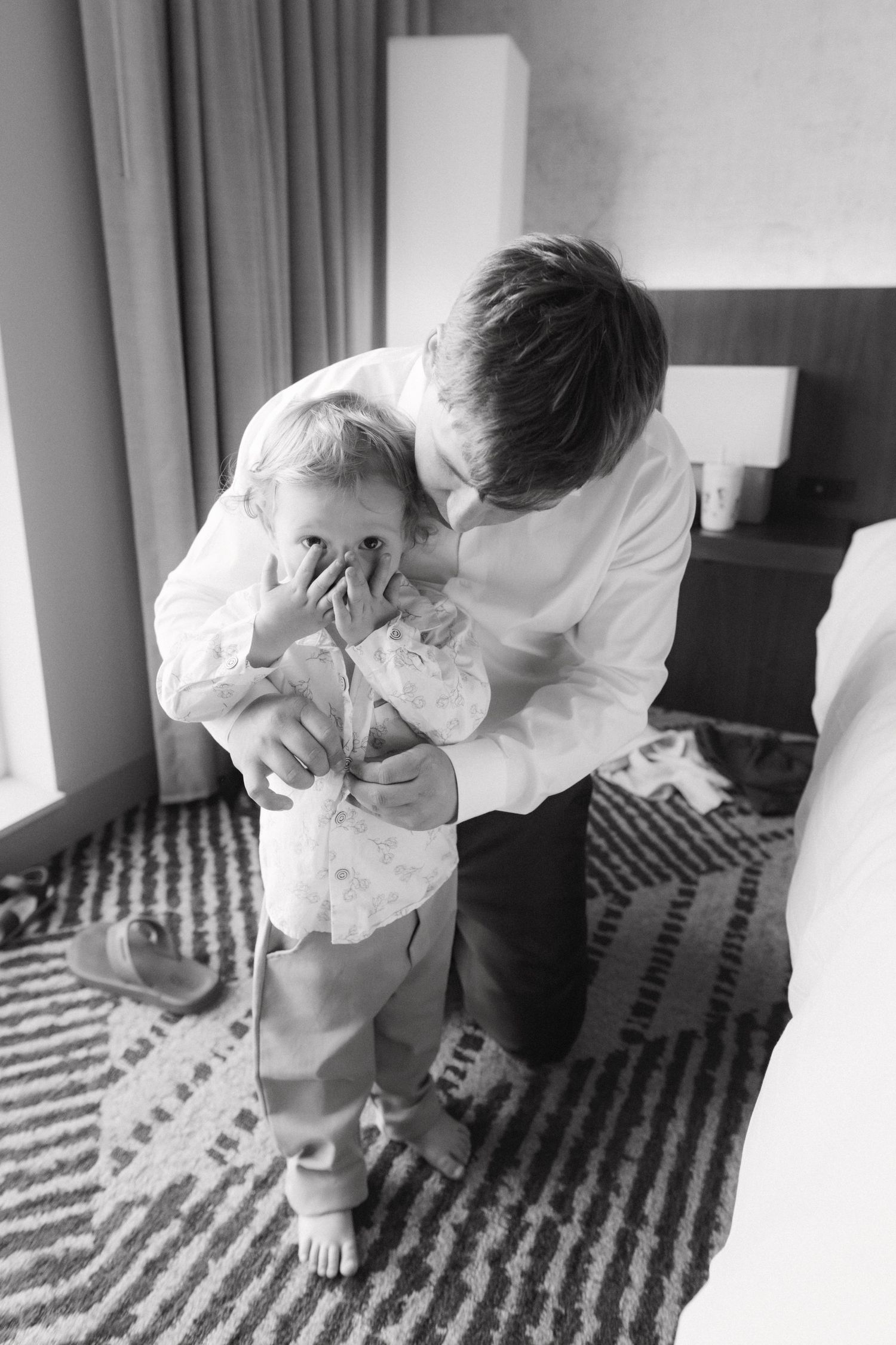 Black and white photo of an older boy helping dress a toddler in a hotel room, both wearing formal white shirts.
