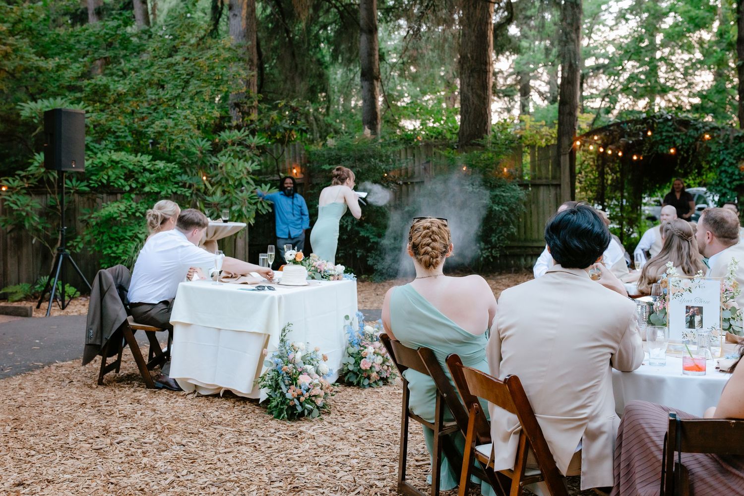 Outdoor wedding reception dinner with guests seated at tables amid trees and string lights.