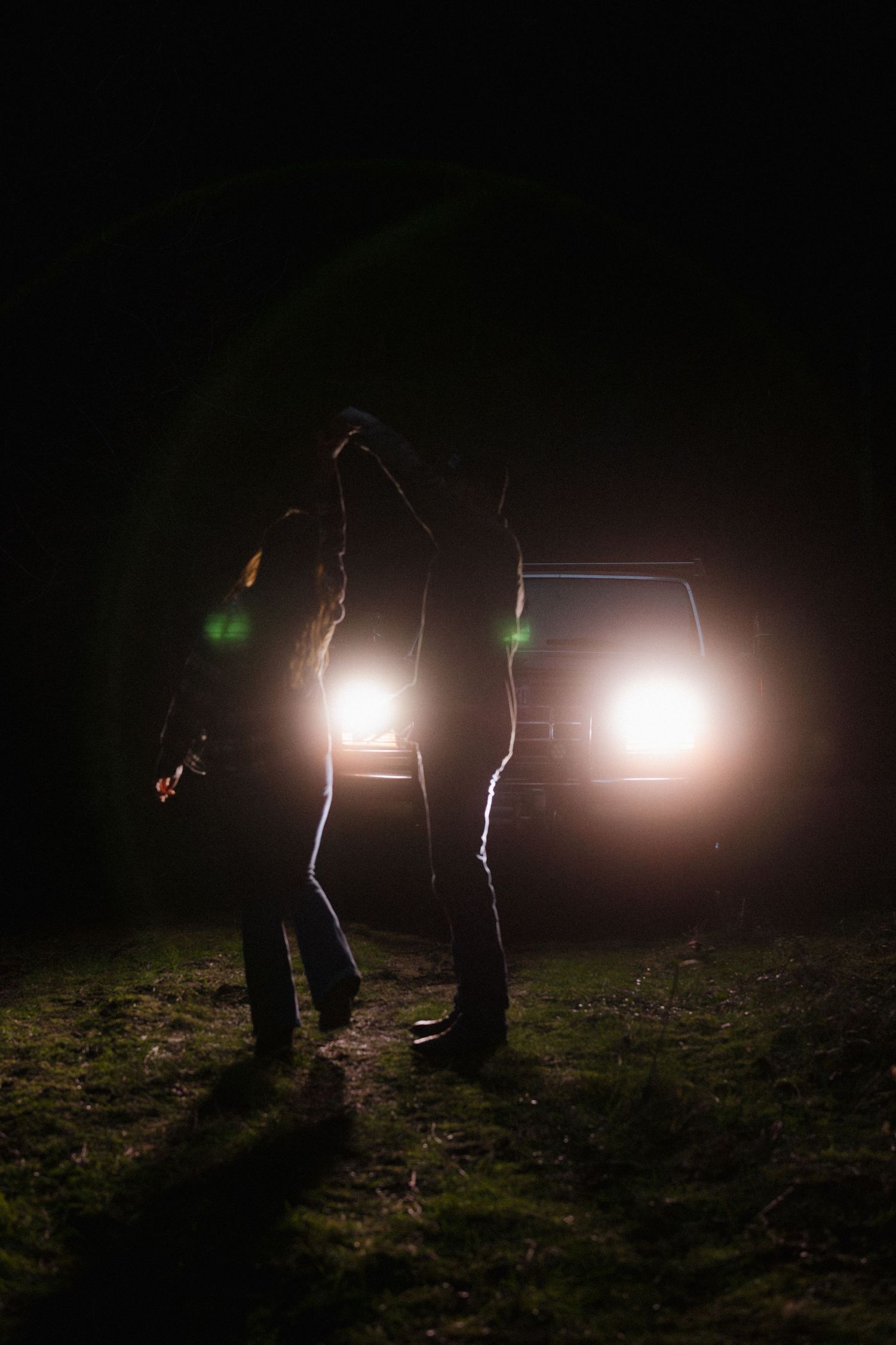 Silhouettes of figures illuminated by car headlights in dark field at night.