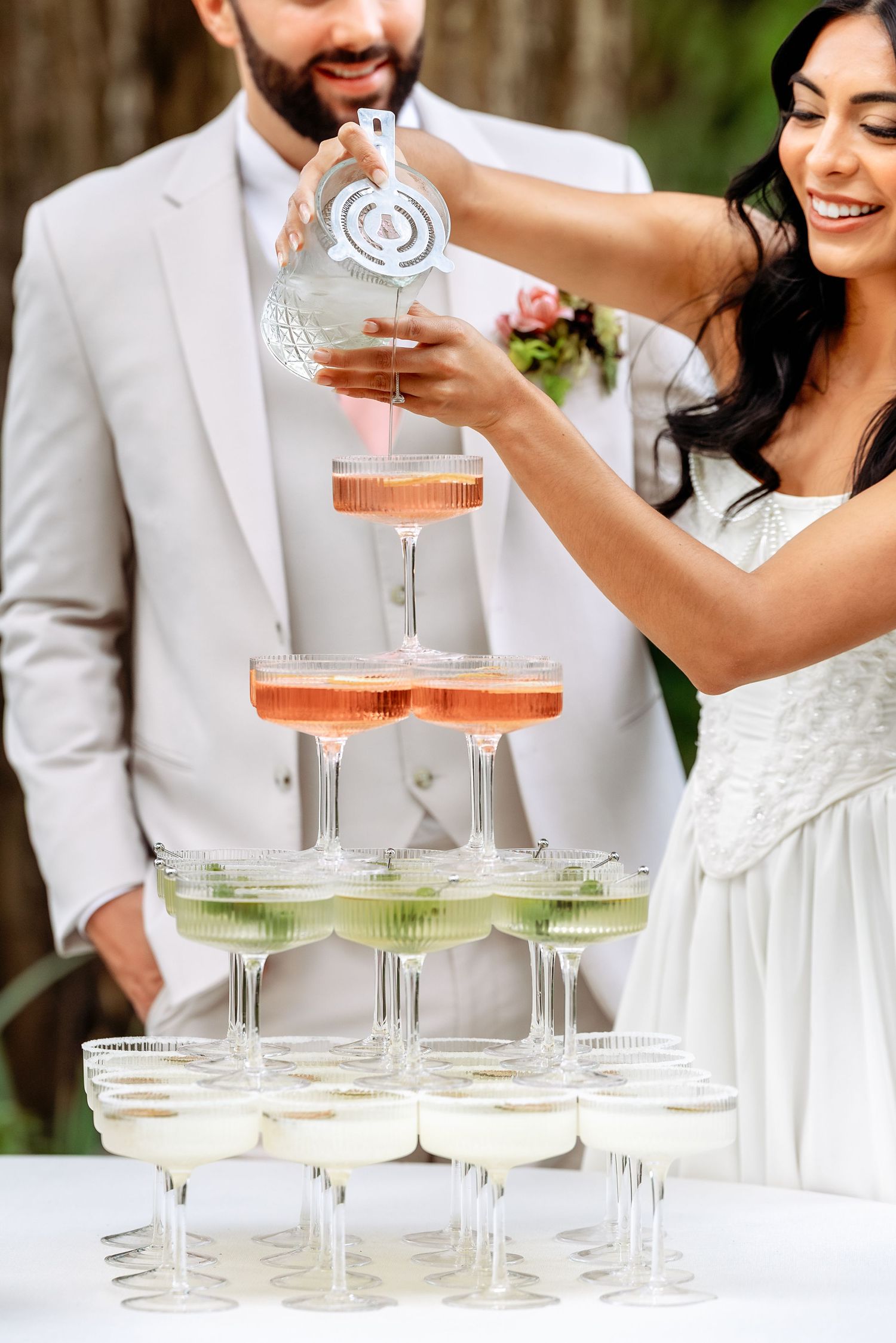 A couple pours champagne into a tower of vintage-style coupe glasses at their wedding celebration.