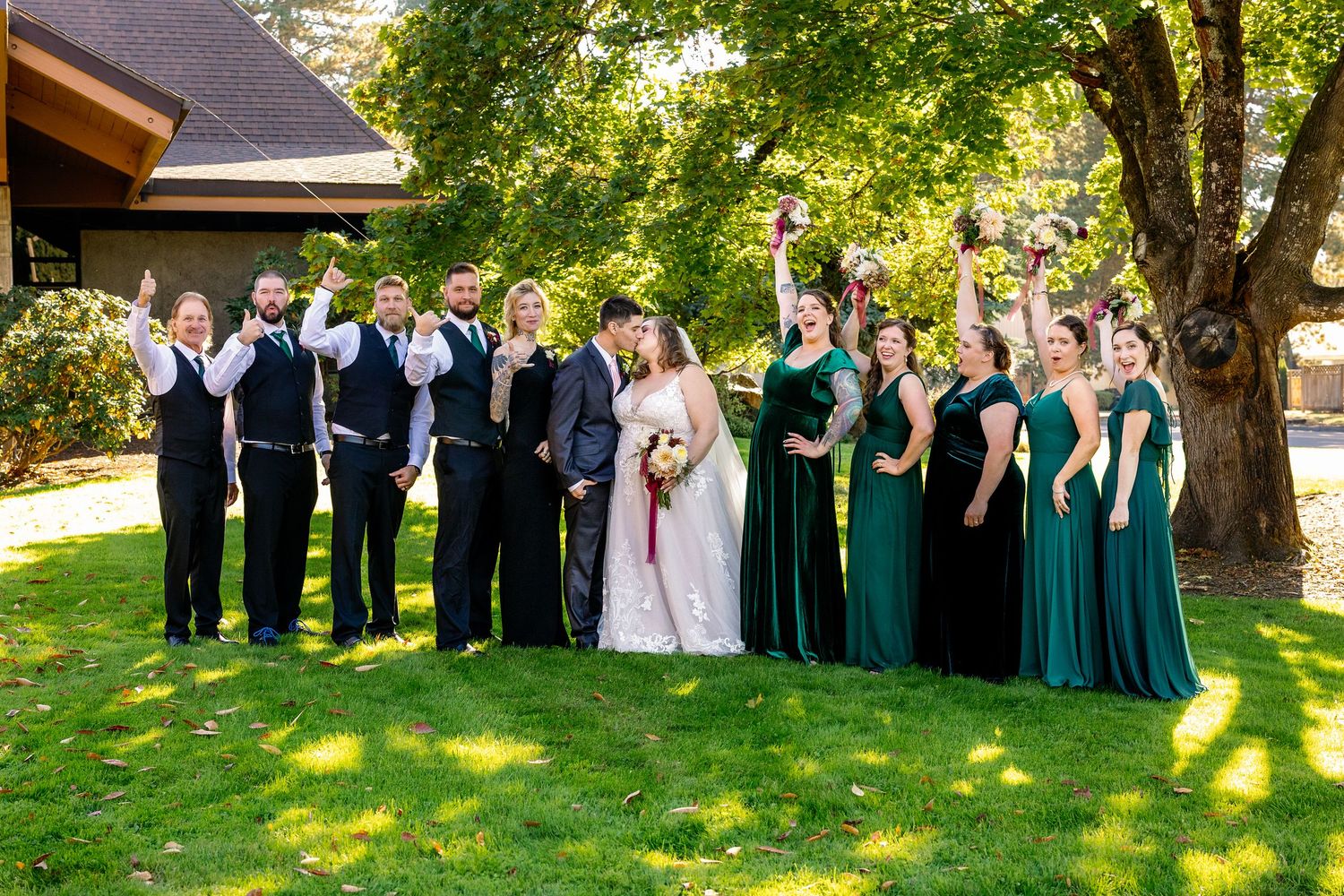 Wedding party in formal attire poses together on a sunny lawn with green trees in the background.