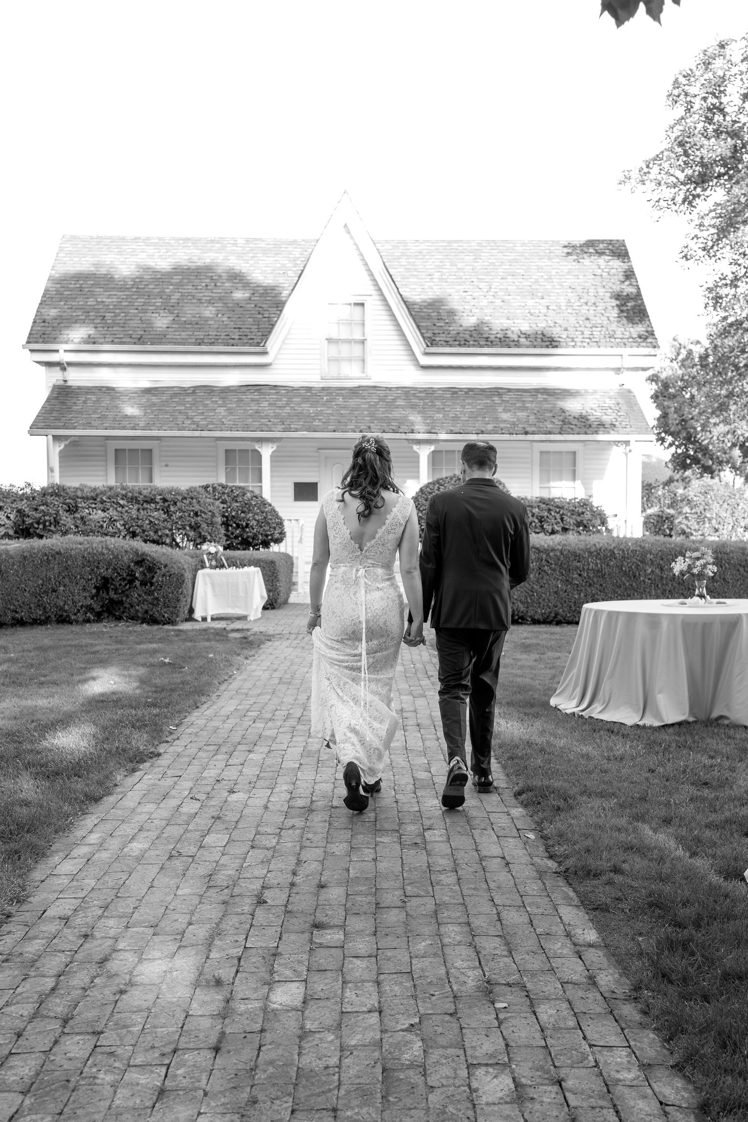 A couple walks away from the camera along a brick pathway with white buildings in the background.