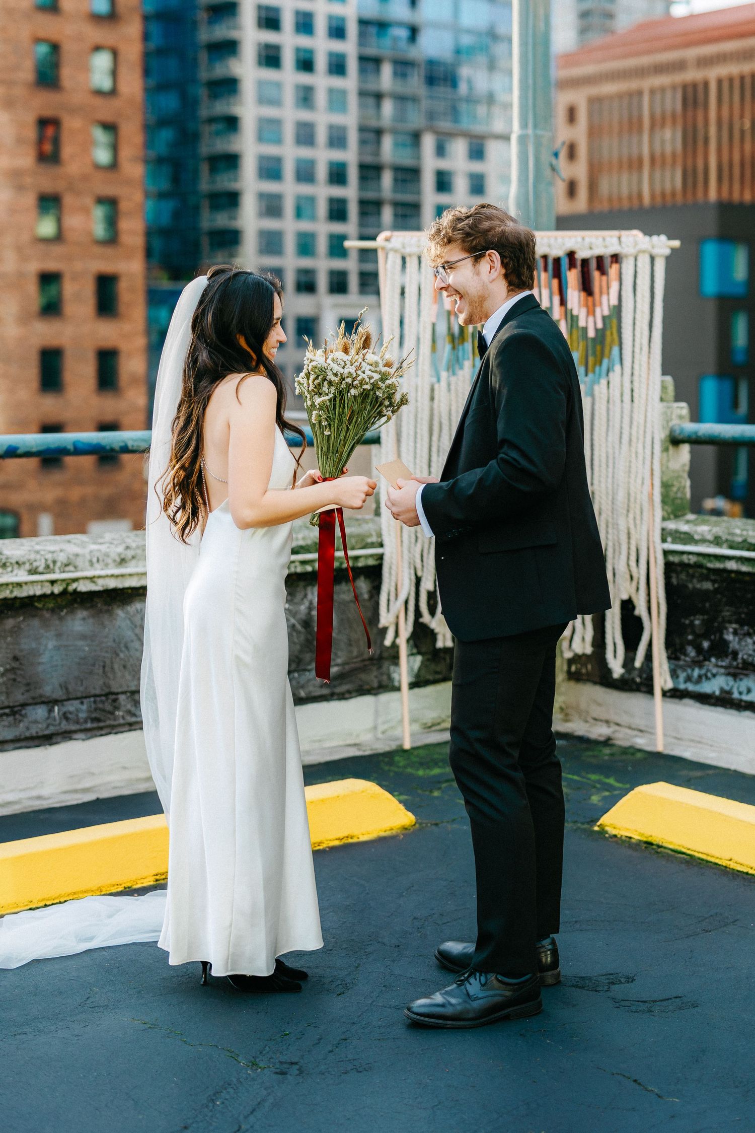 Couple in formal wear sharing romantic moment on urban rooftop with macrame backdrop.