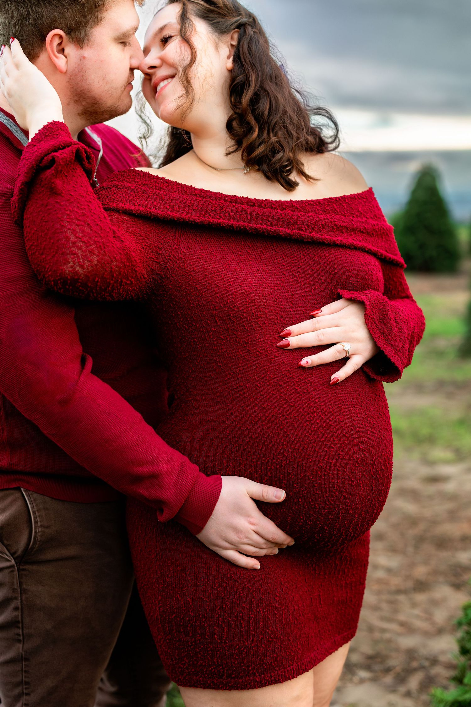 A romantic maternity photo showing a couple embracing outdoors, with the person wearing a cozy red sweater dress.