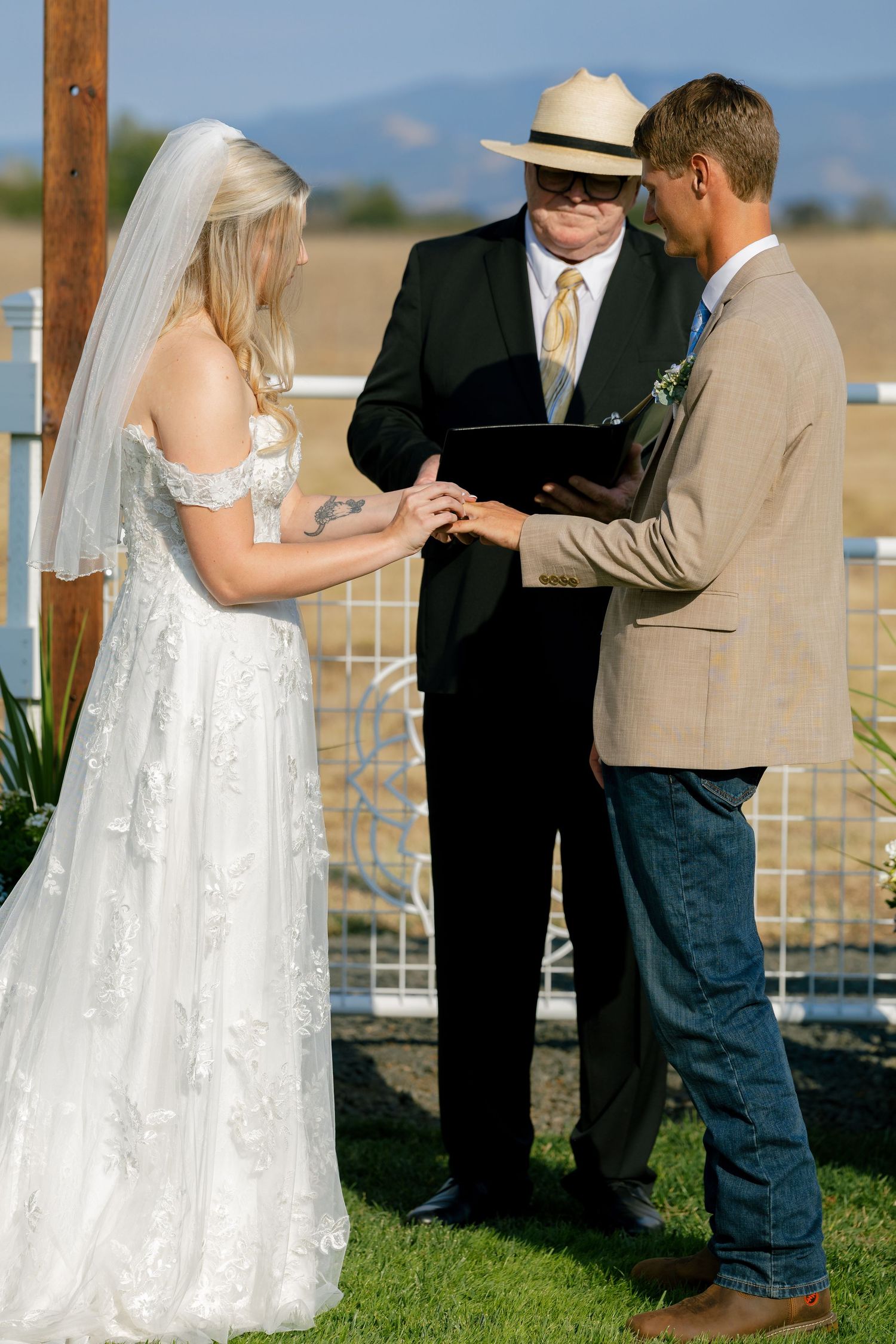 Wedding ceremony exchange of rings under wooden arch with mountain backdrop.