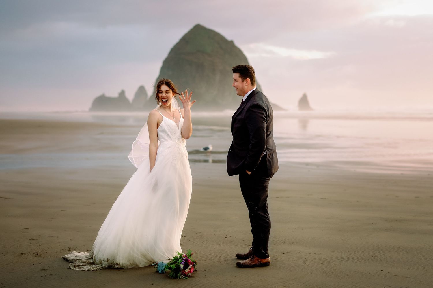 A couple shares a romantic moment on a misty beach with dramatic coastal rock formations in the background.