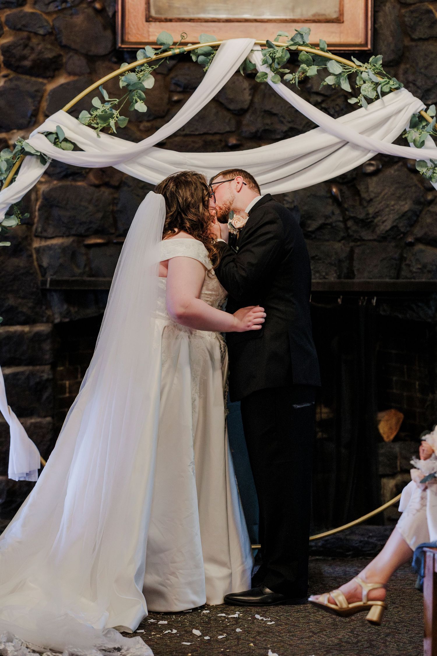 Wedding ceremony under white draped fabric and greenery arch against stone wall backdrop.