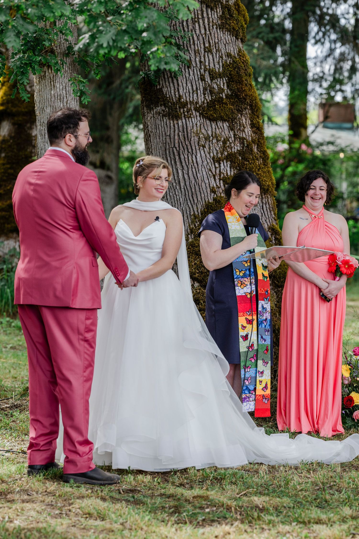 Wedding party in coral and white dresses gathers under large trees during outdoor summer celebration.