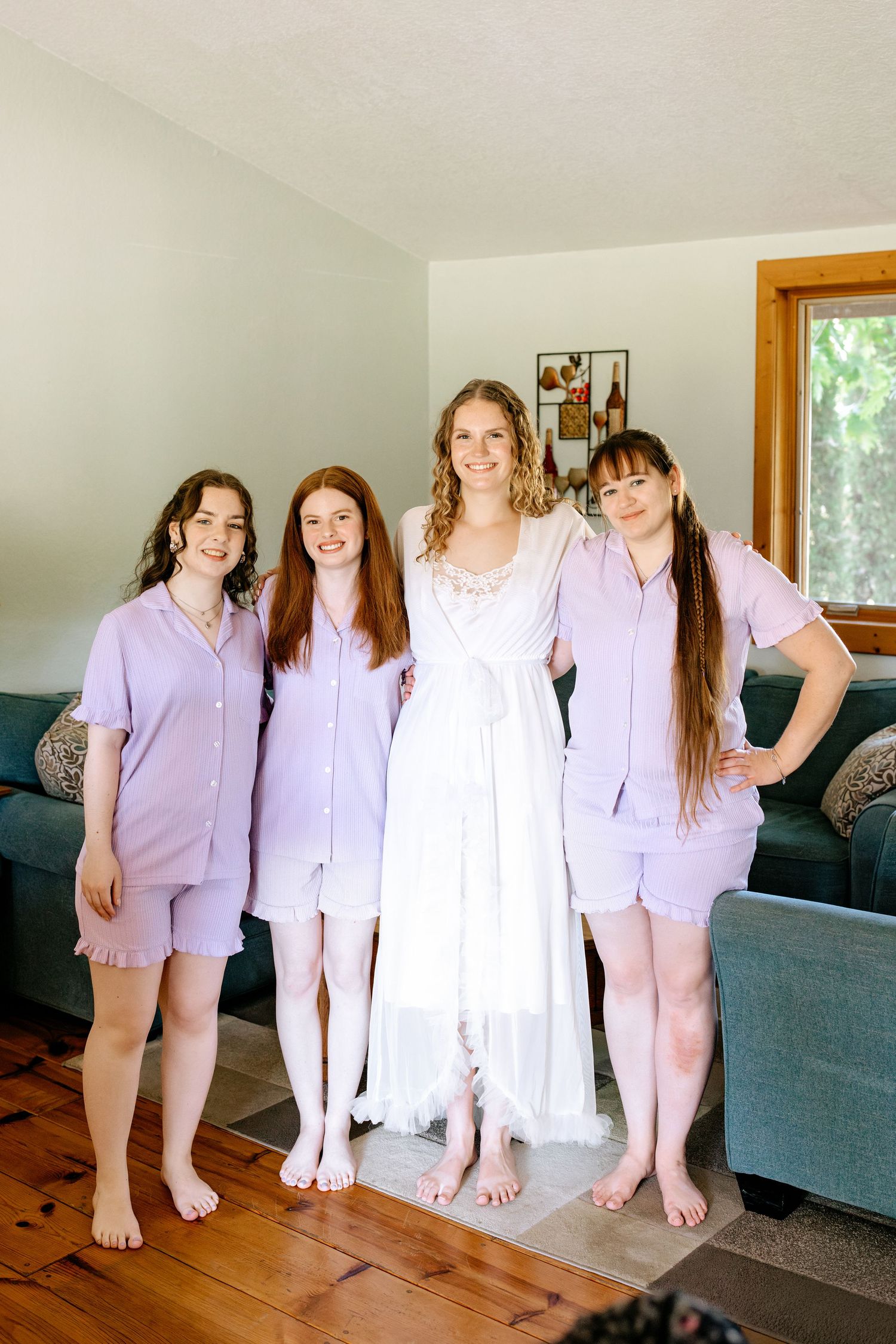 Four women pose indoors, three in matching lavender pajamas beside a bride in a white wedding robe.