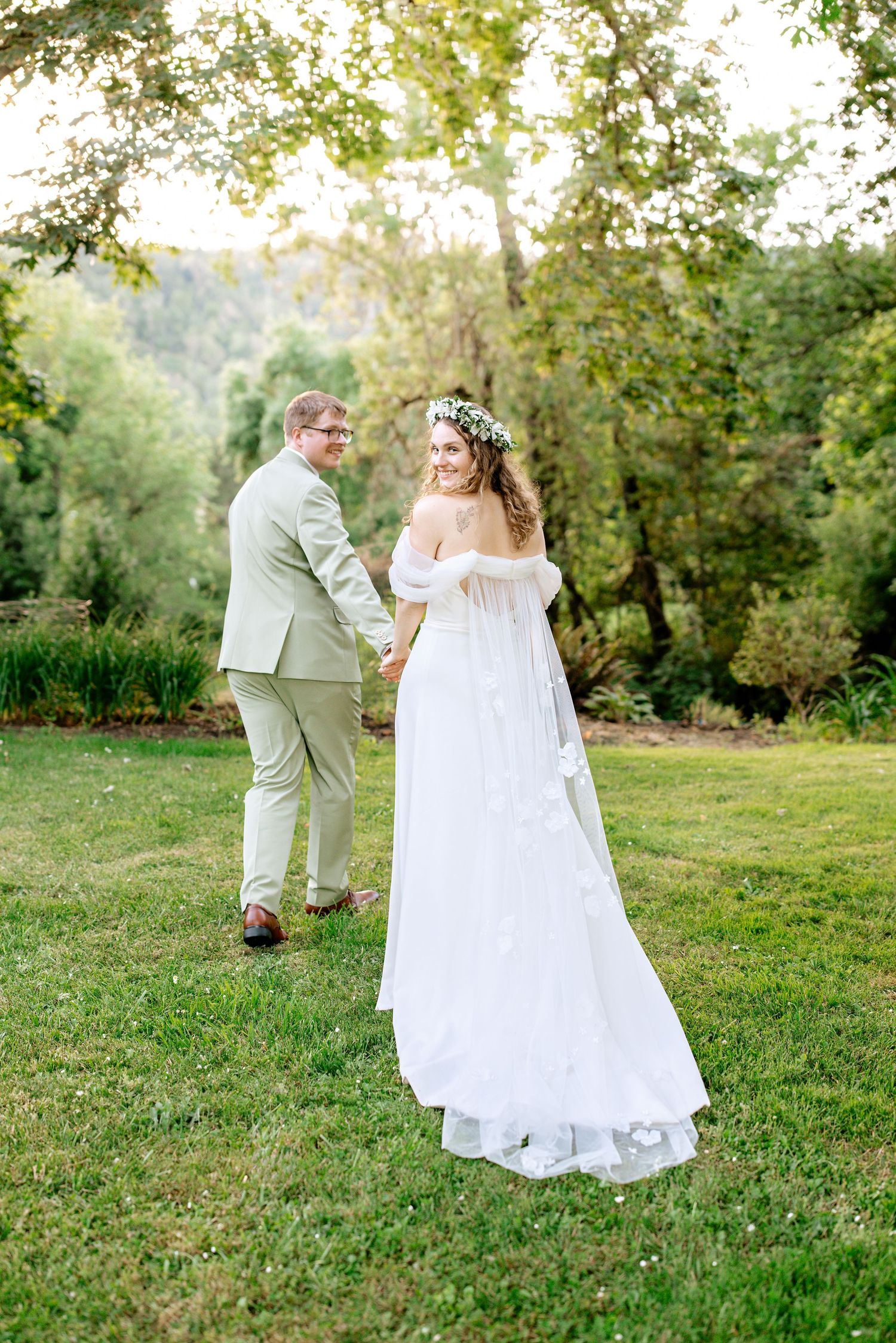 Bride in floral crown glances back smiling as groom leads her by hand across sunlit garden lawn.
