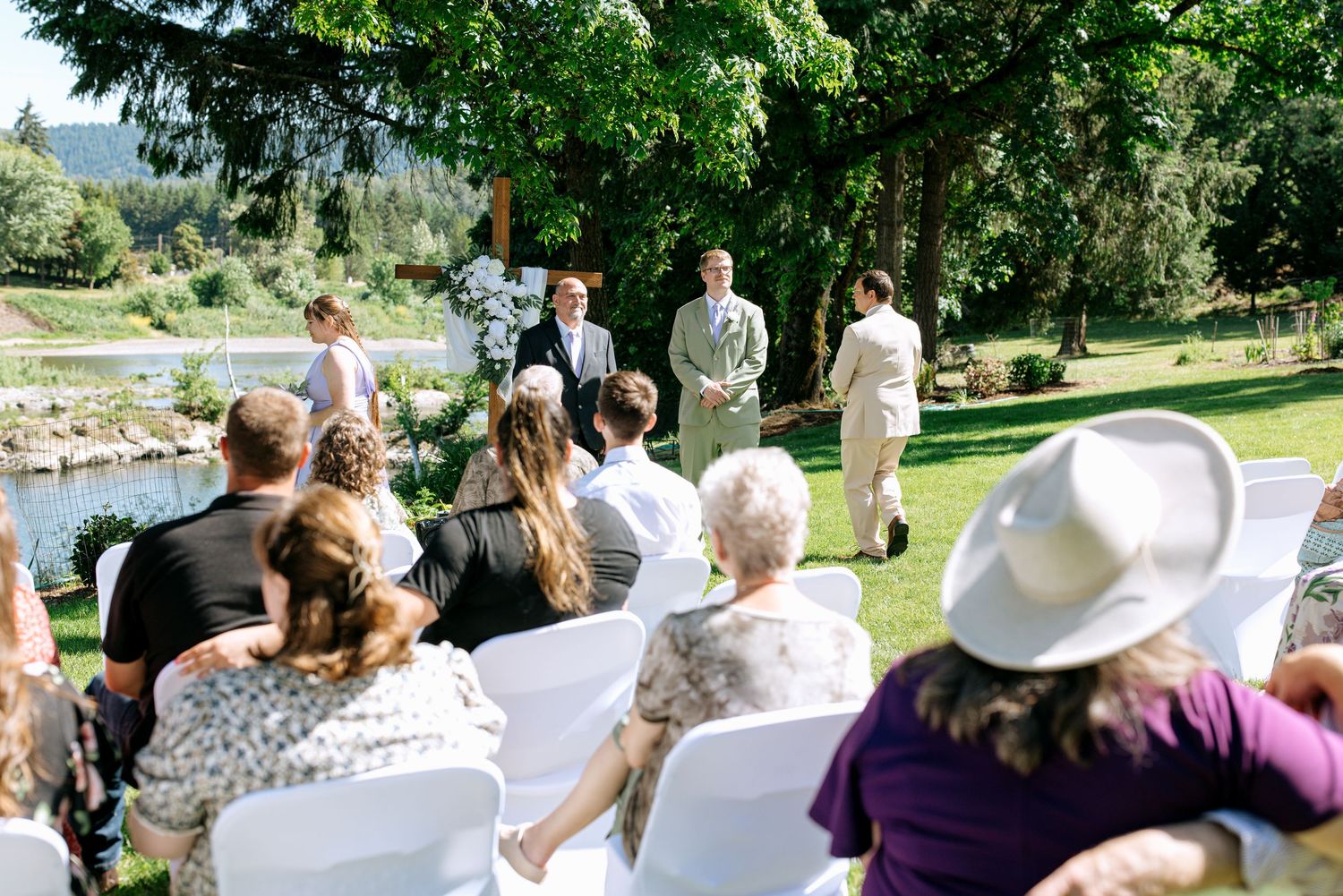 Outdoor wedding ceremony on green lawn with guests seated in white chairs facing couple at floral arch by riverside.