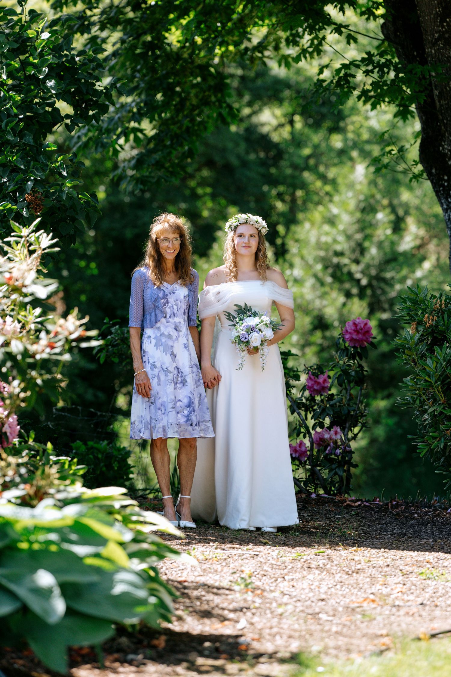 Bride in white off-shoulder gown with floral crown holds bouquet beside woman in lavender dress in lush garden.