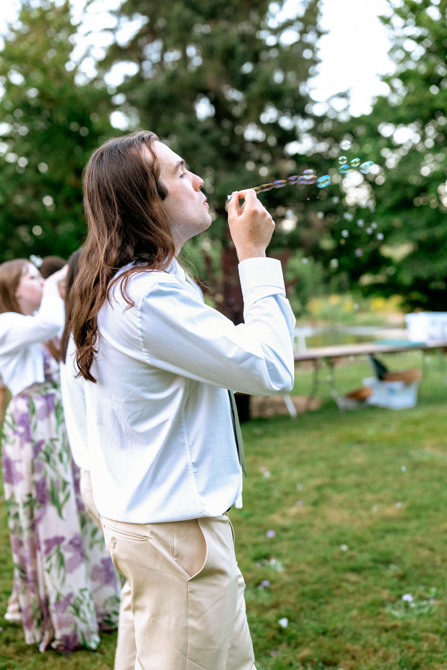 Young man in white shirt blowing bubbles at an outdoor wedding reception.