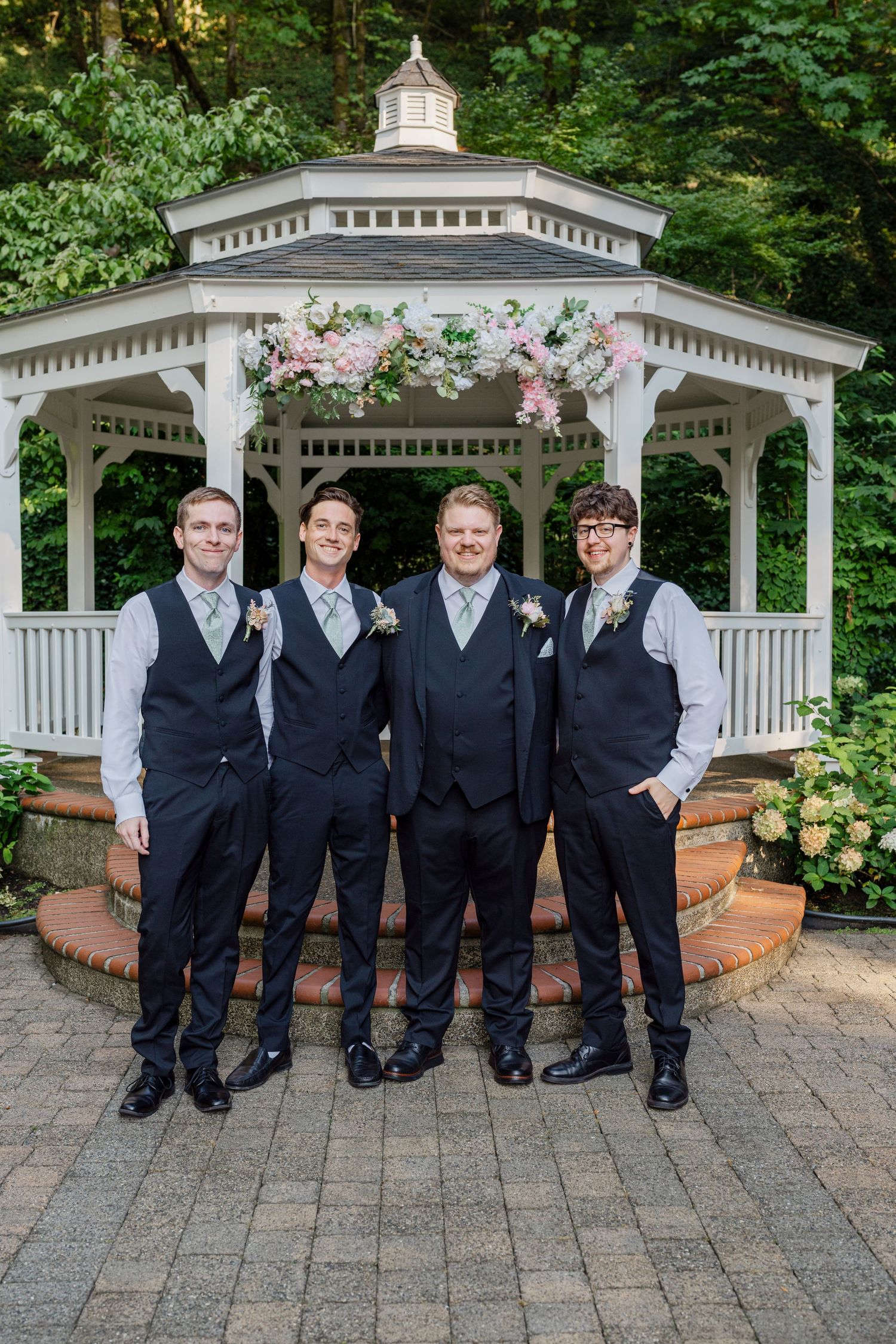 Four groomsmen in navy vests pose together in front of a white floral-adorned gazebo outdoors.