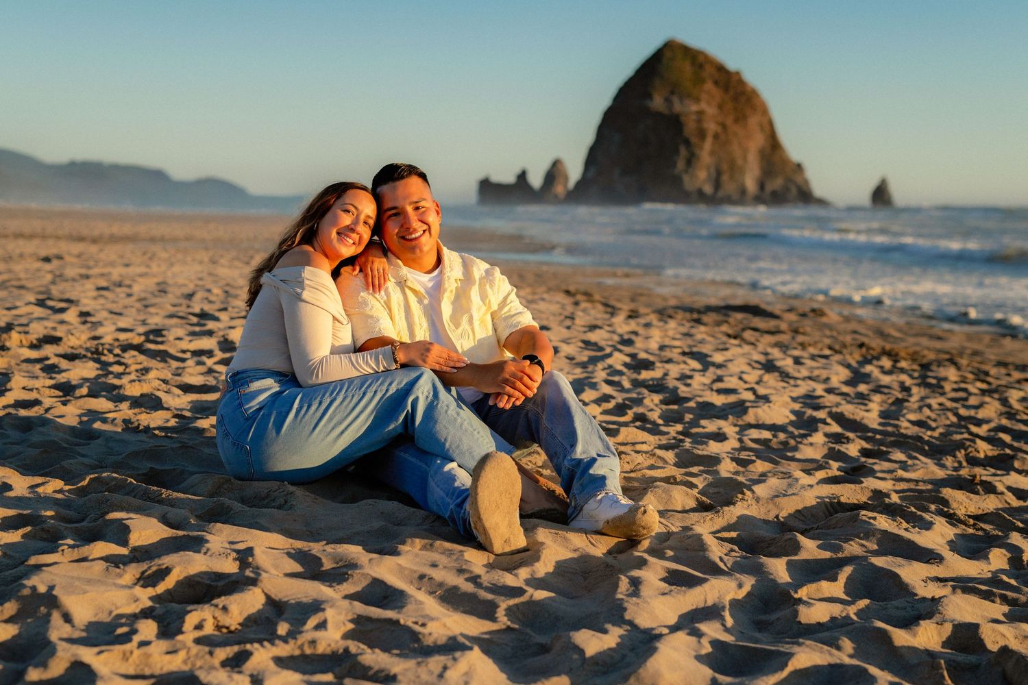 Couple sitting together on sandy beach at golden hour with Haystack Rock in background at Cannon Beach Oregon.