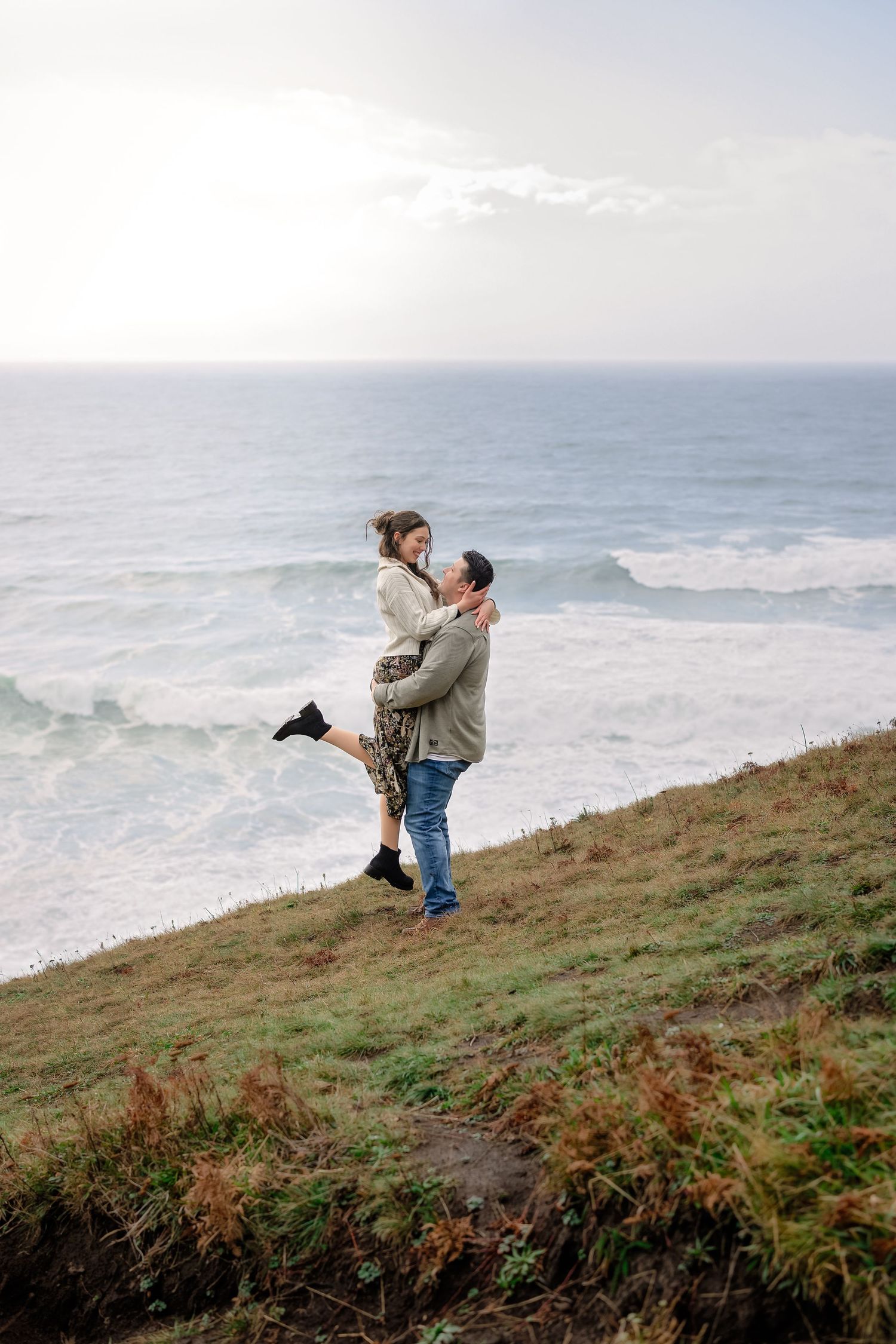 Couple shares tender moments on coastal clifftop with dramatic ocean views and misty horizon.