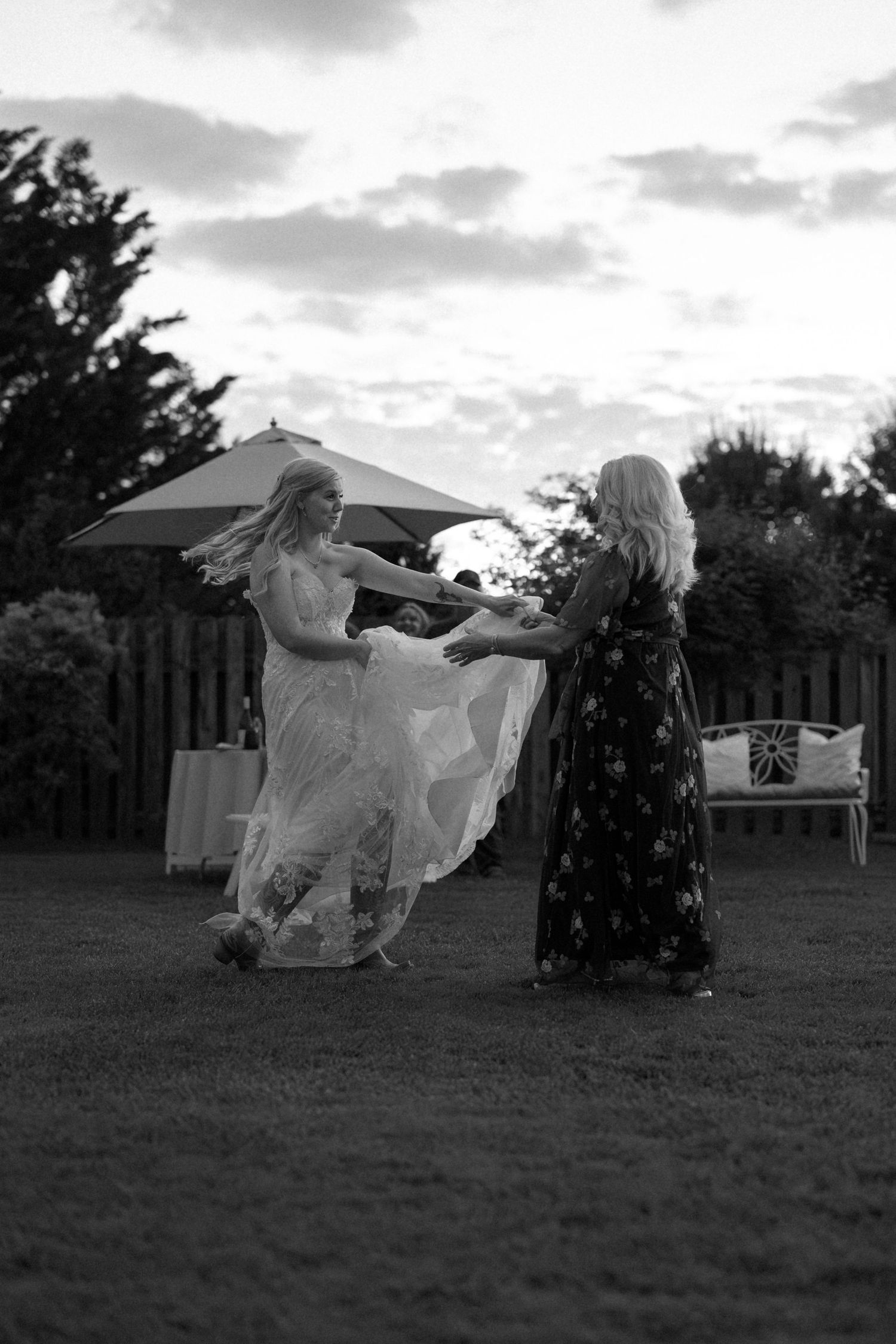 Two figures dance together at an outdoor wedding reception as the sun sets behind them in black and white.