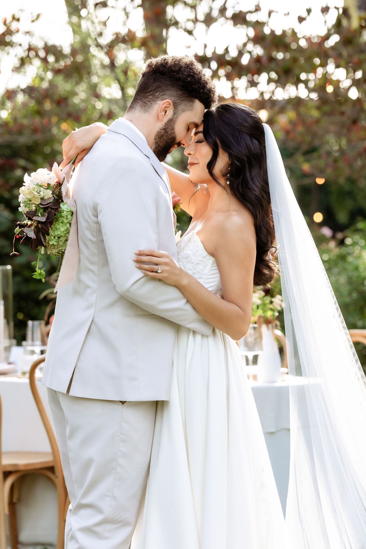 A romantic wedding couple embraces outdoors with the bride holding a bouquet against a soft bokeh background.