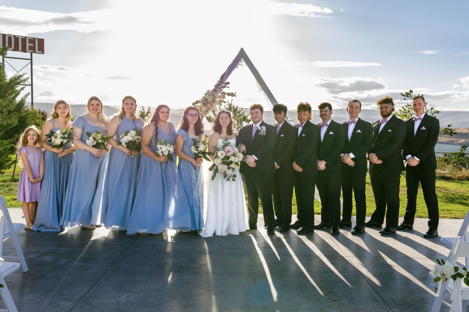 Wedding party lineup with bridesmaids in blue dresses and groomsmen in dark suits against scenic backdrop.