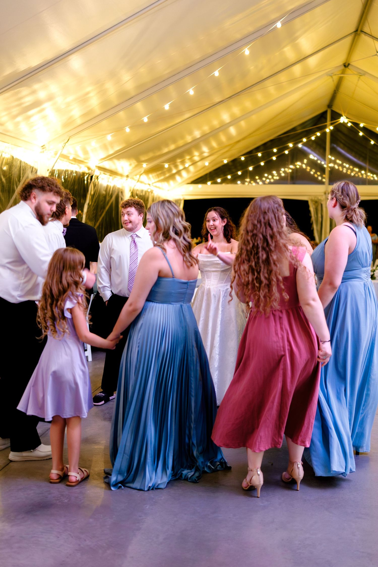 Wedding guests in blue and pink dresses dance together under a tent with string lights.