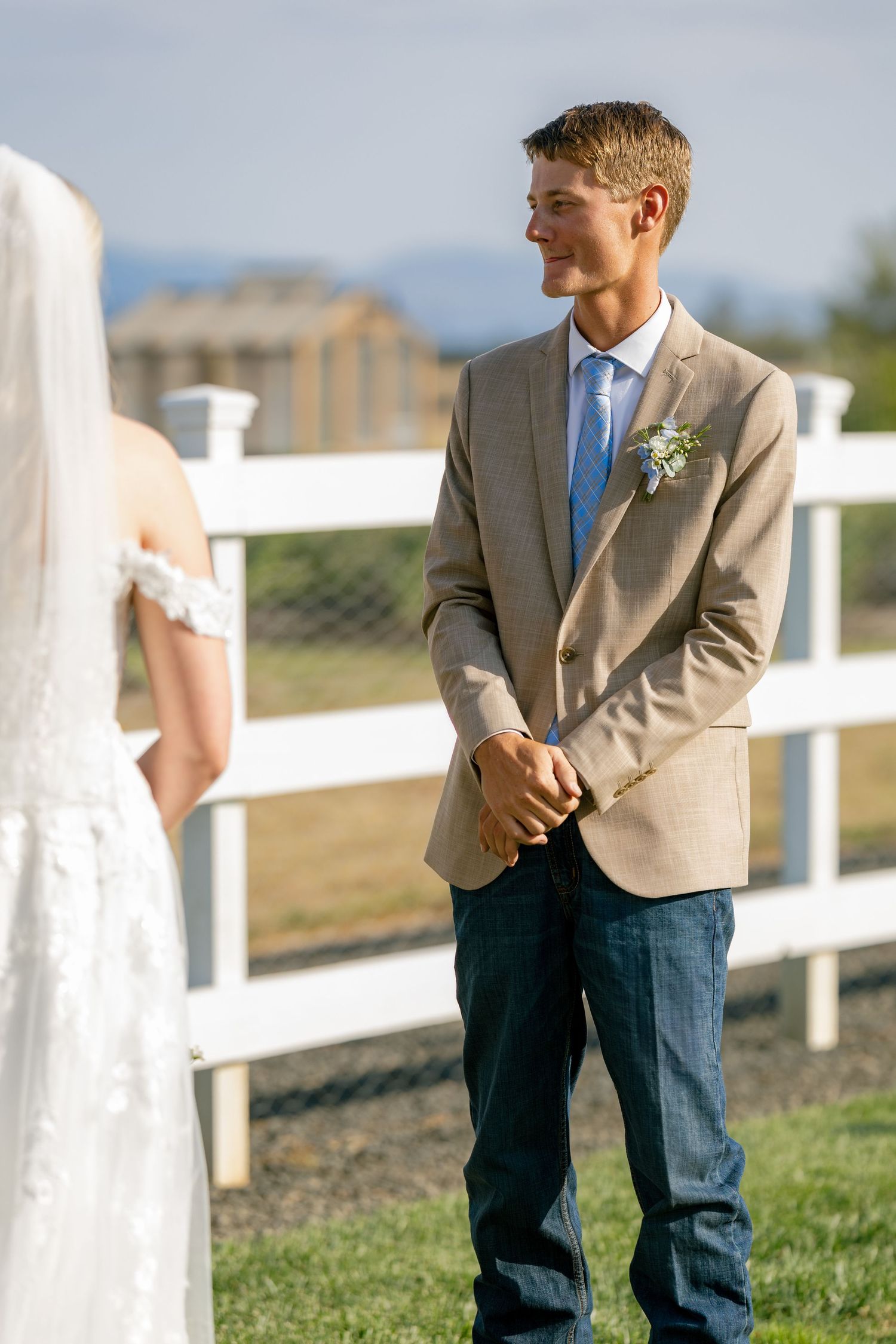 Person in a tan suit jacket and jeans standing by a white fence at an outdoor venue.
