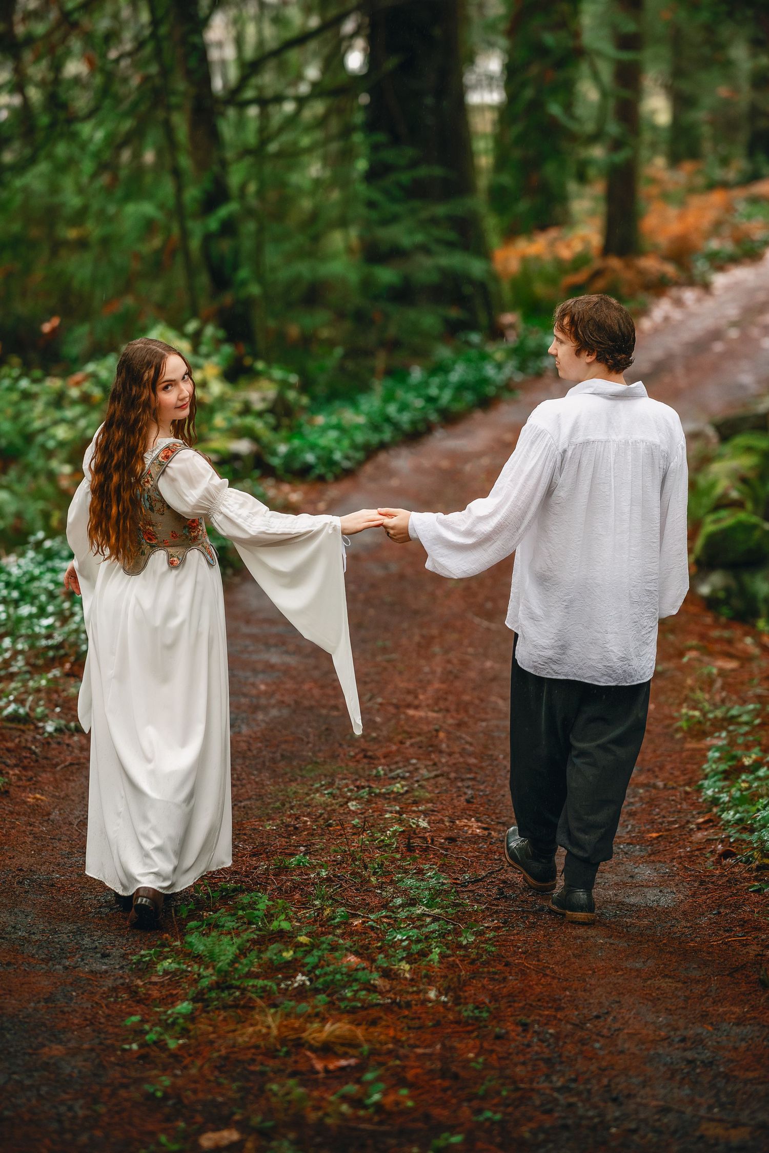 Two people in white and black attire walk hand in hand down a forest path.