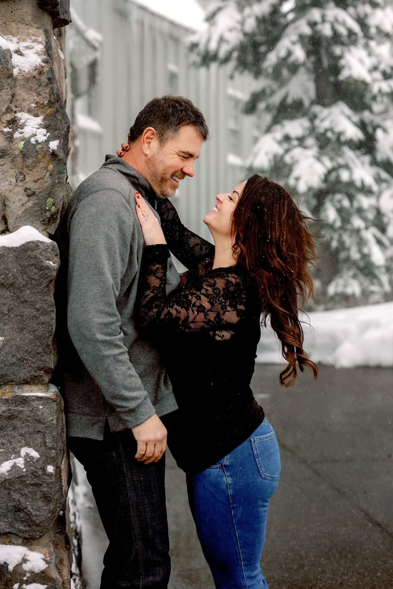 A couple embraces and laughs together during a winter photoshoot with snow-covered trees in the background.