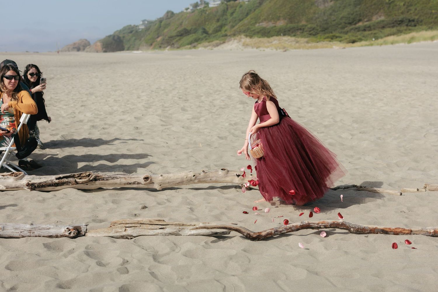 People in formal attire sitting on driftwood at a beach during sunset.