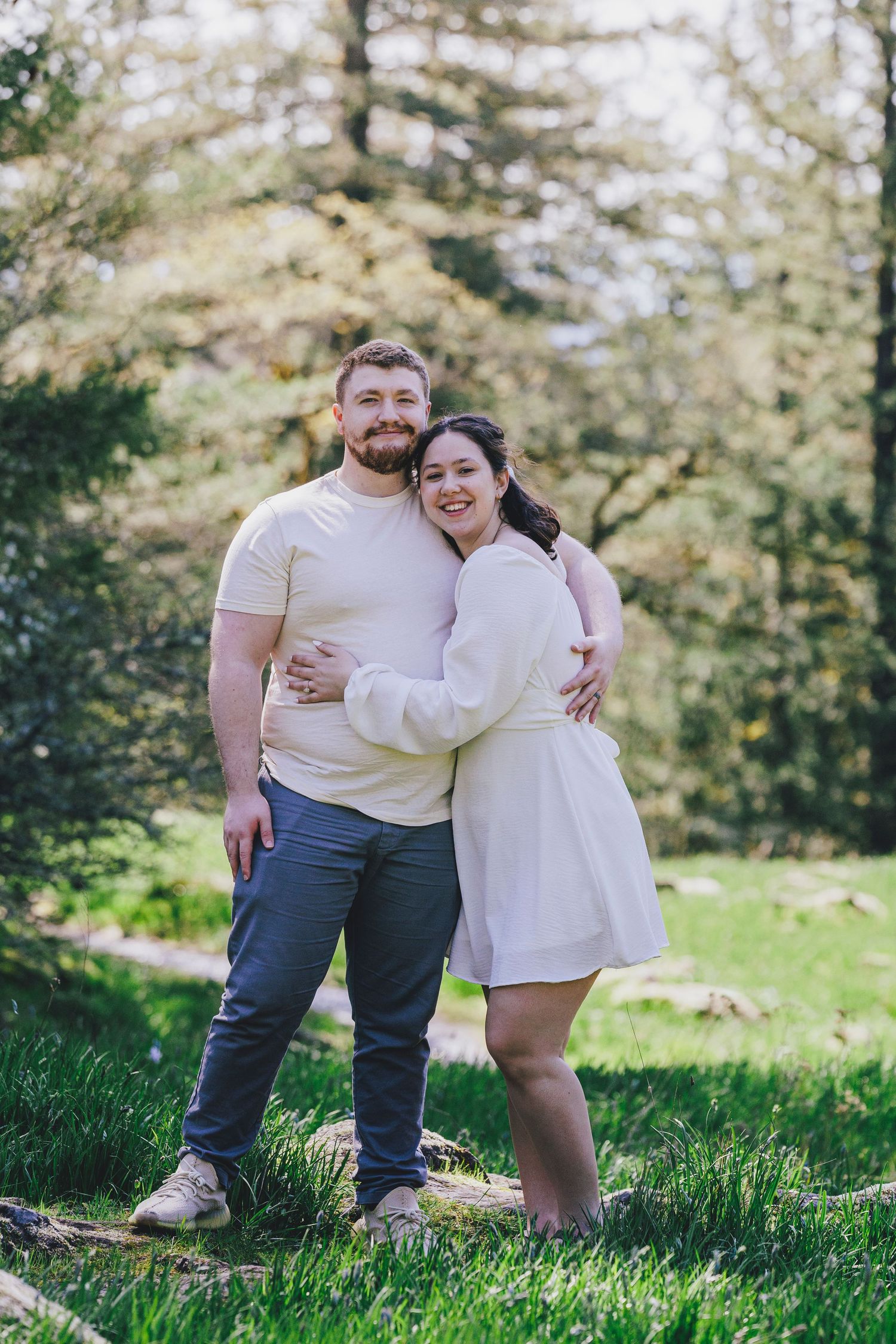 A romantic couple wearing coordinated white outfits embraces while standing in a sunny forest clearing.