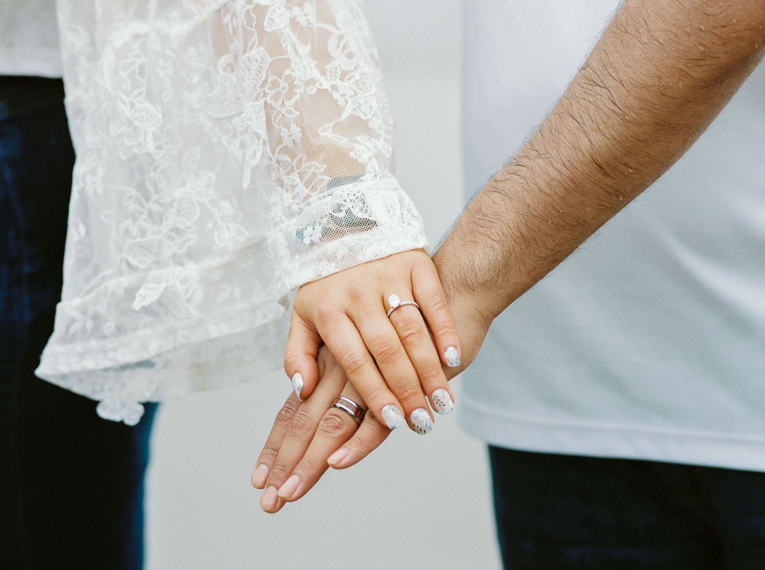 Couple holding hands showing wedding and engagement rings, bride in white lace dress sleeve, romantic wedding photo.