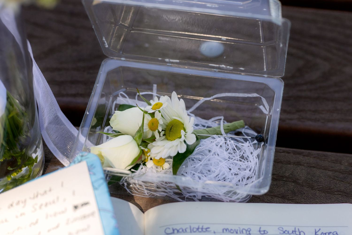 White flowers and greenery in a clear plastic box with handwritten notes nearby.