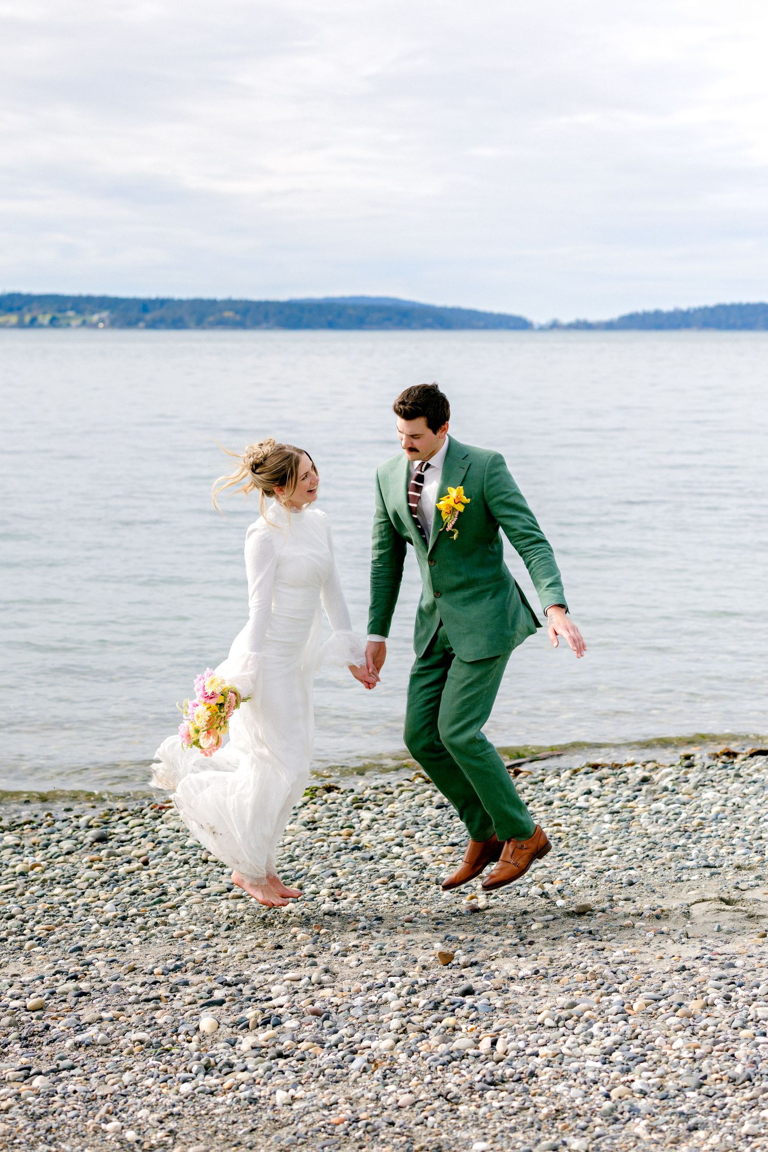 Bride and groom hold hands laughing while walking on pebble beach by the water on wedding day.